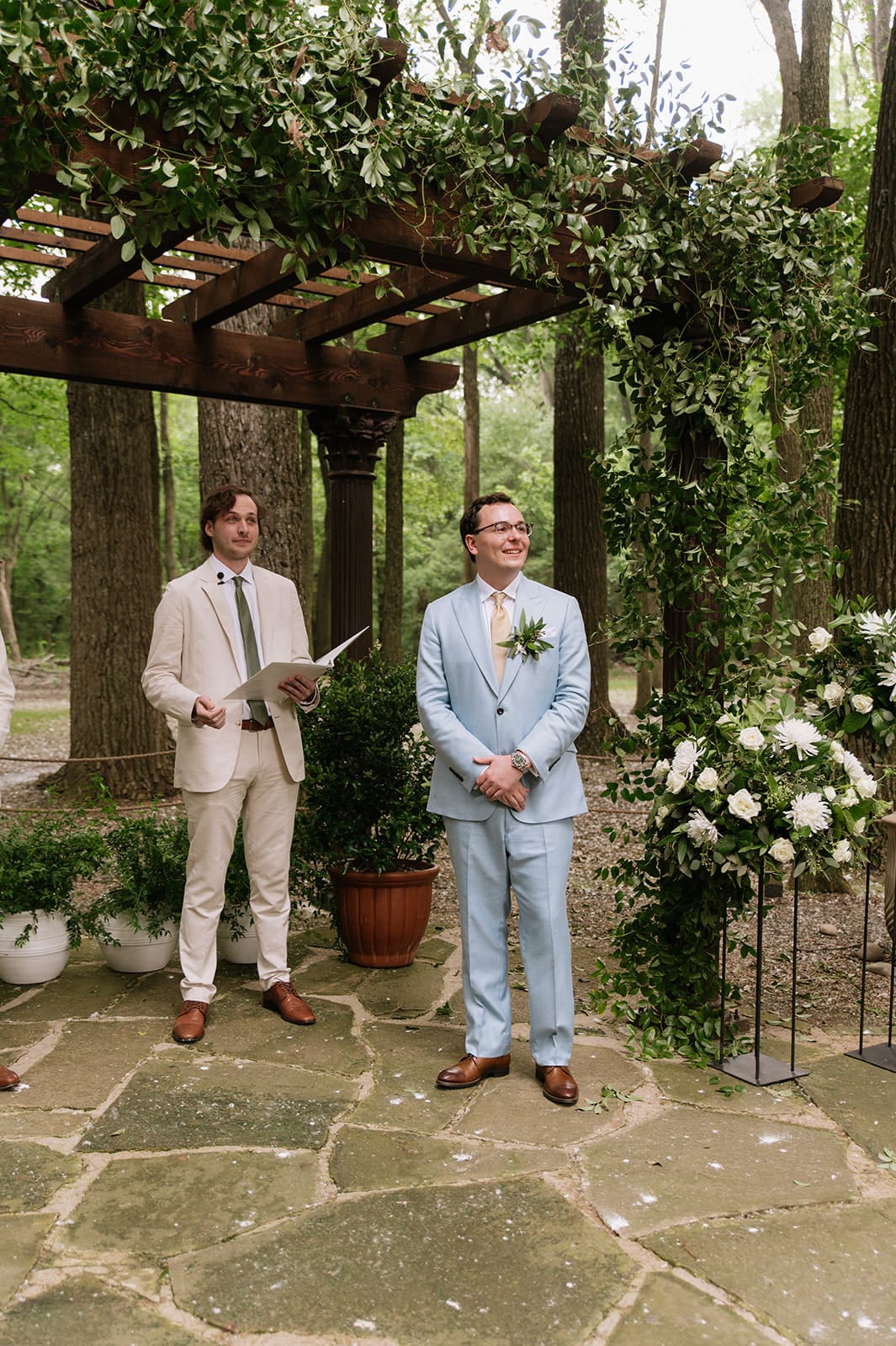 Two men stand under a pergola decorated with greenery and flowers; one holds a book and the other, in a blue suit, stands with hands clasped, both outdoors on a stone patio at The Springs Waxahachie