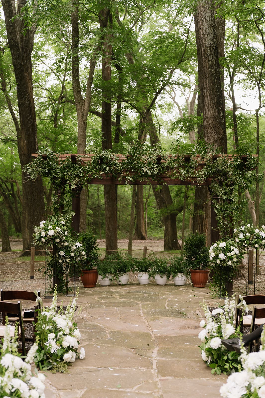 An outdoor wedding ceremony setup in a forest features a floral arch, potted plants, and white flowers lining the stone aisle, with trees in the background at The Springs Waxahachie