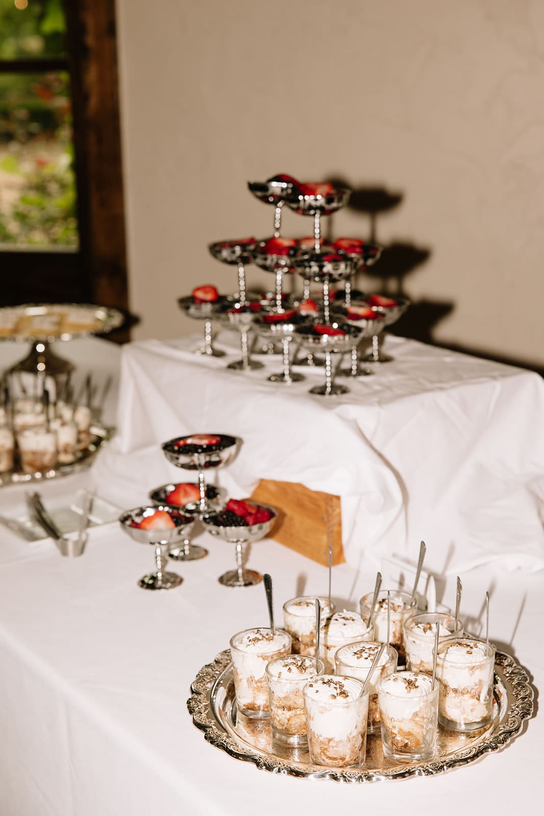 A dessert table with layered parfaits in glasses on a silver tray and fruit-topped desserts displayed on tiered stands, all set on a white tablecloth.