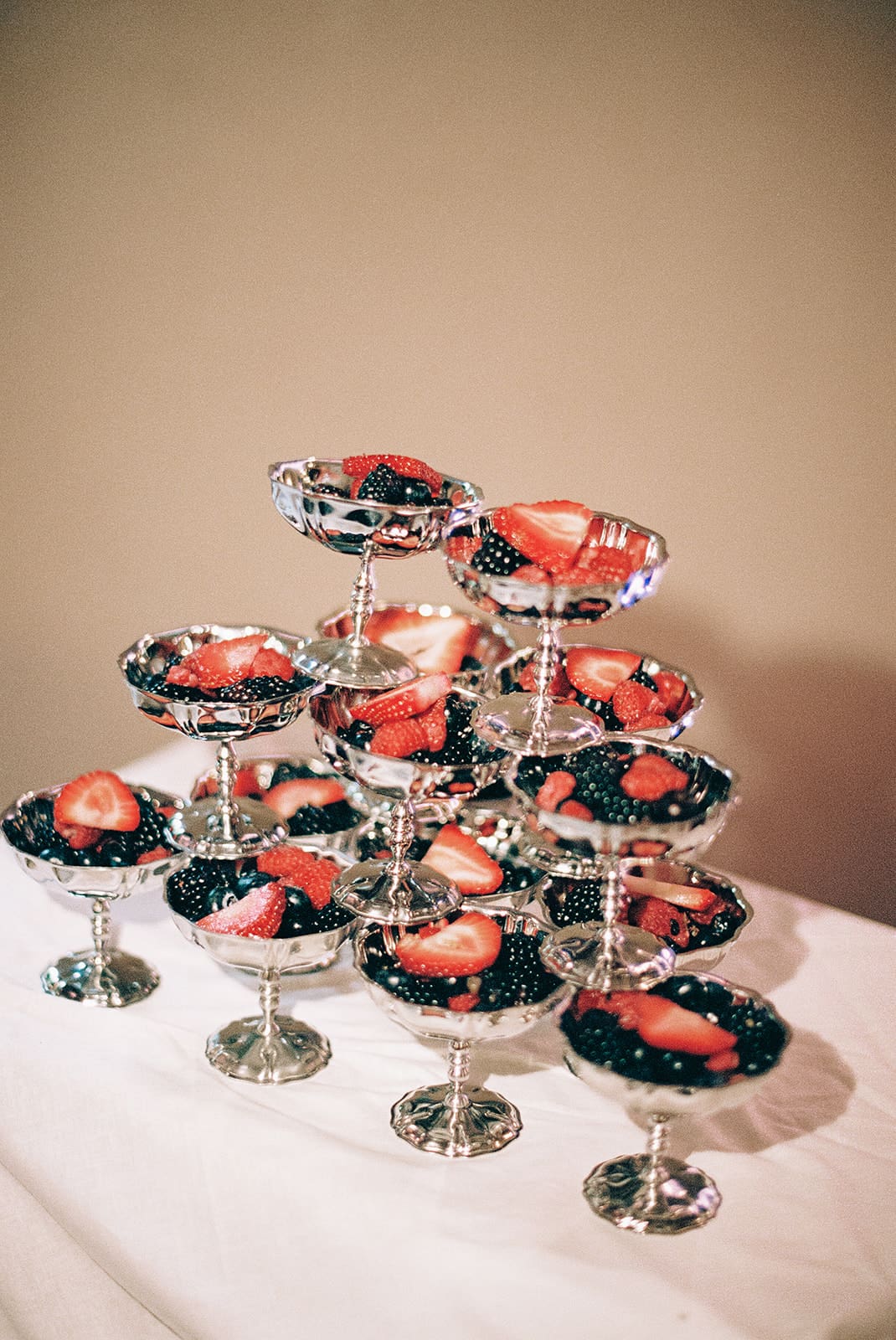 A dessert table with layered parfaits in glasses on a silver tray and fruit-topped desserts displayed on tiered stands, all set on a white tablecloth.