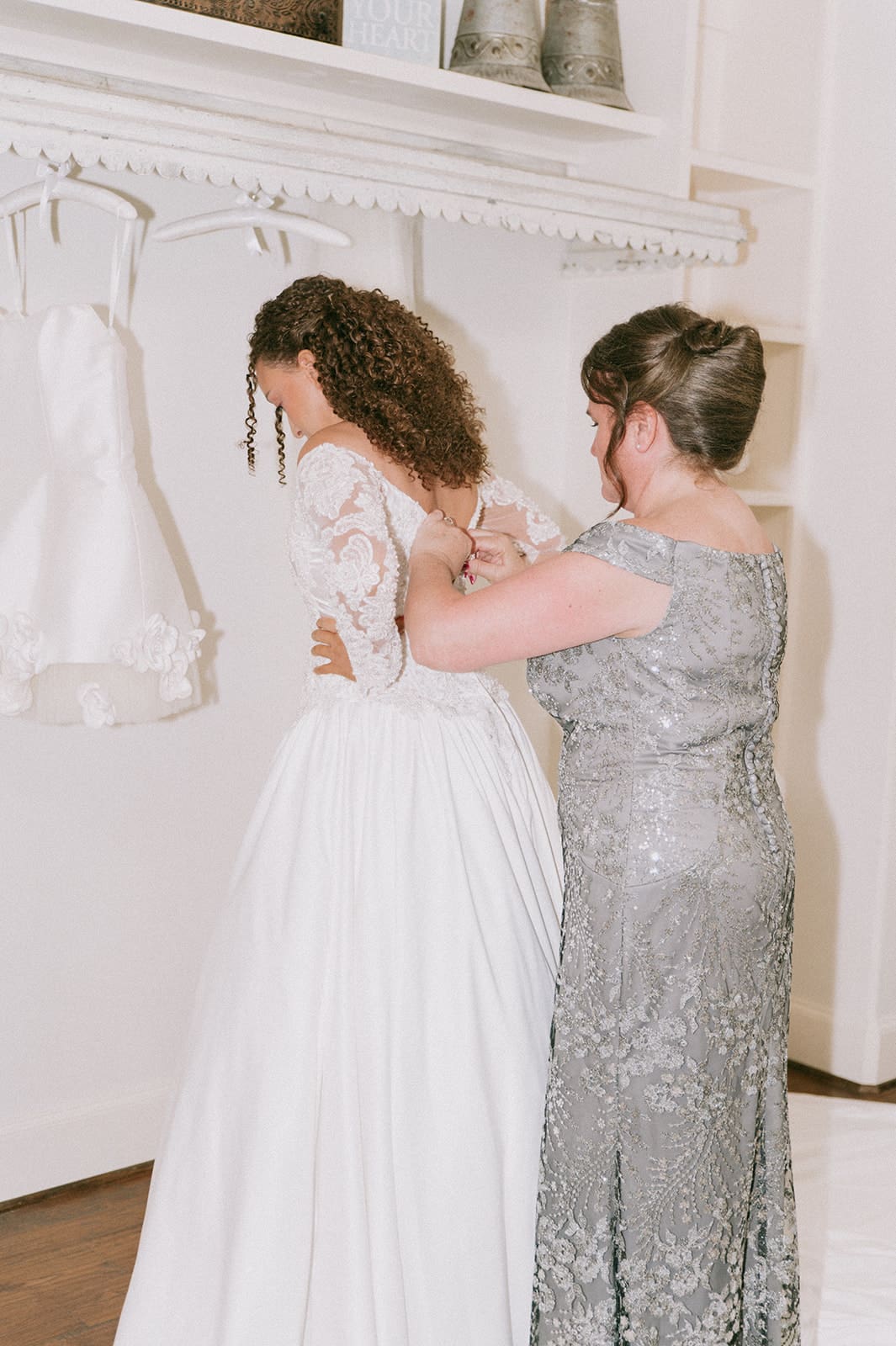 A woman in a silver dress helps another woman in a white wedding gown adjust her dress 