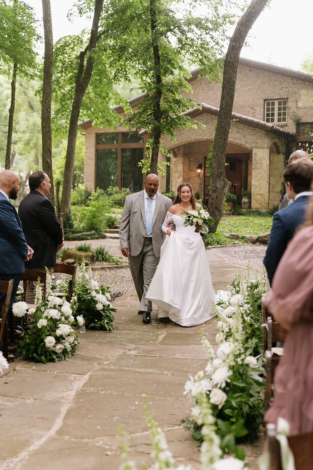 A bride in a white gown, holding a bouquet, walks down an outdoor aisle lined with flowers, accompanied by a man in a light suit. Guests are seated on either side at The Springs Waxahachie