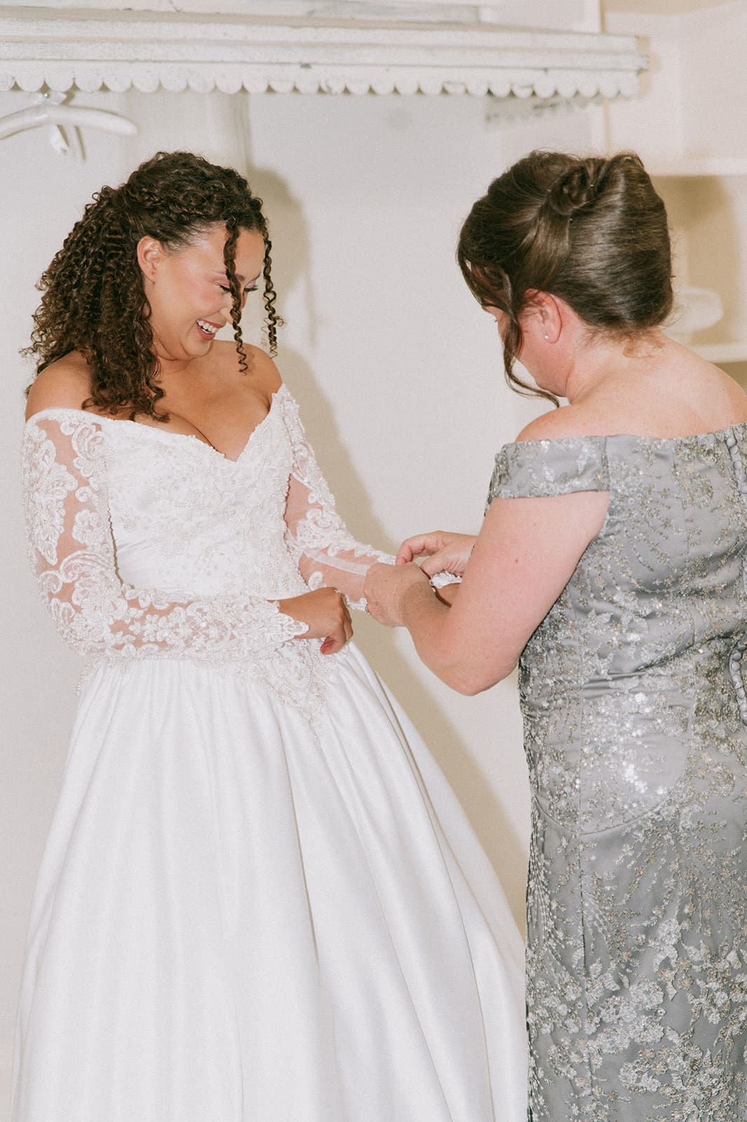 A woman in a silver dress helps another woman in a white wedding gown adjust her dress 