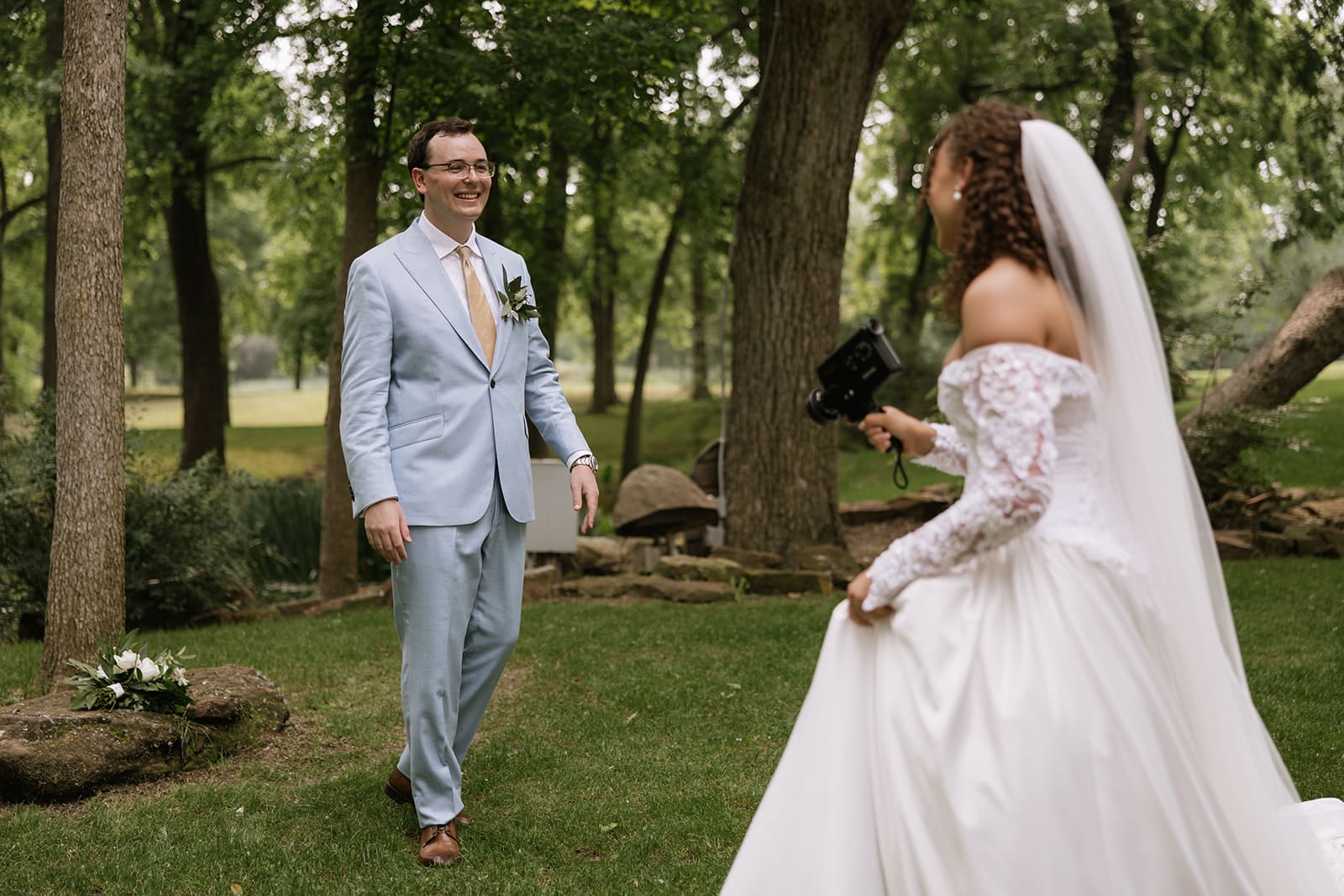 A bride in a white dress and veil approaches a groom in a light blue suit in a wooded outdoor setting, both smiling.