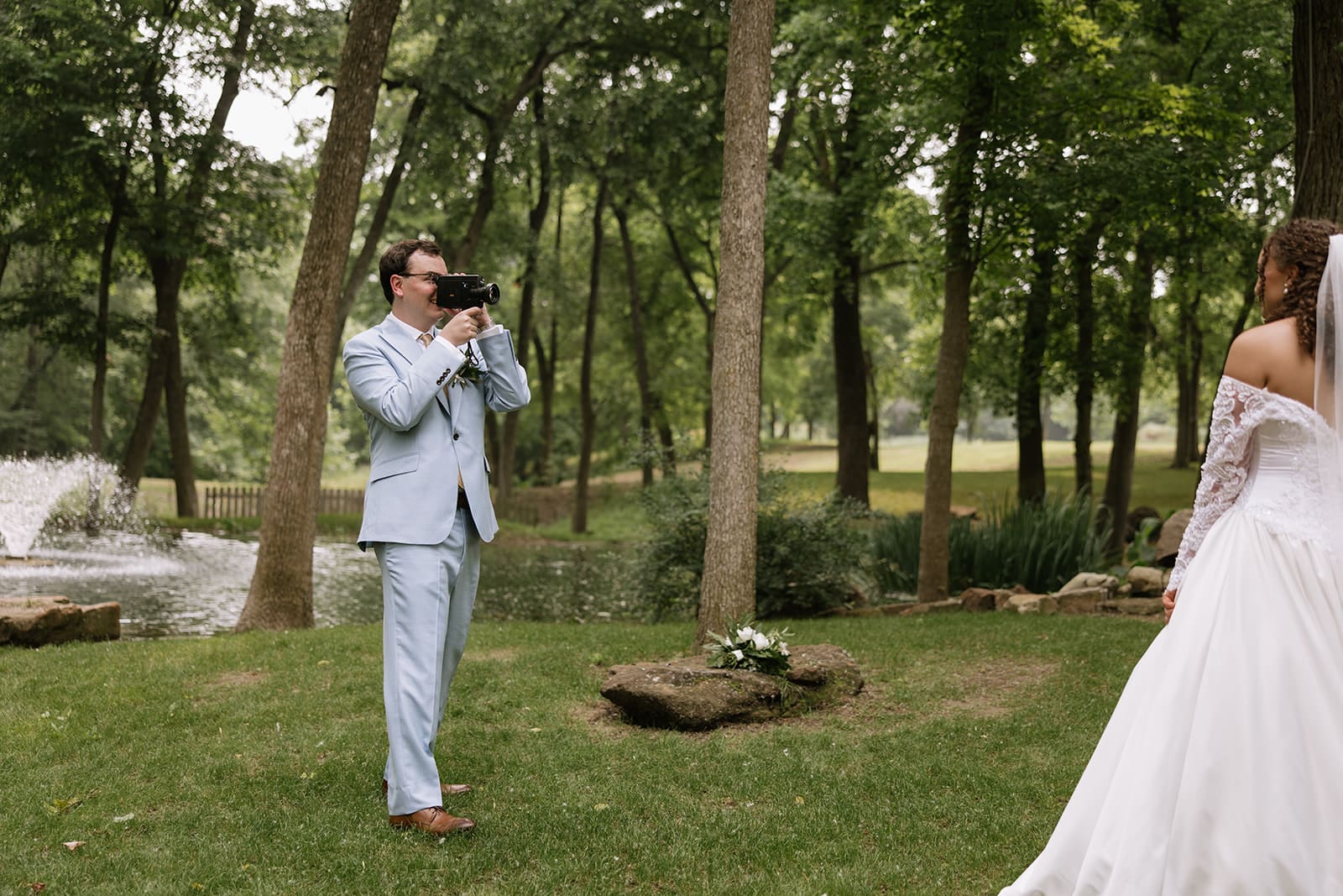 A bride in a white dress and veil approaches a groom in a light blue suit in a wooded outdoor setting, both smiling.