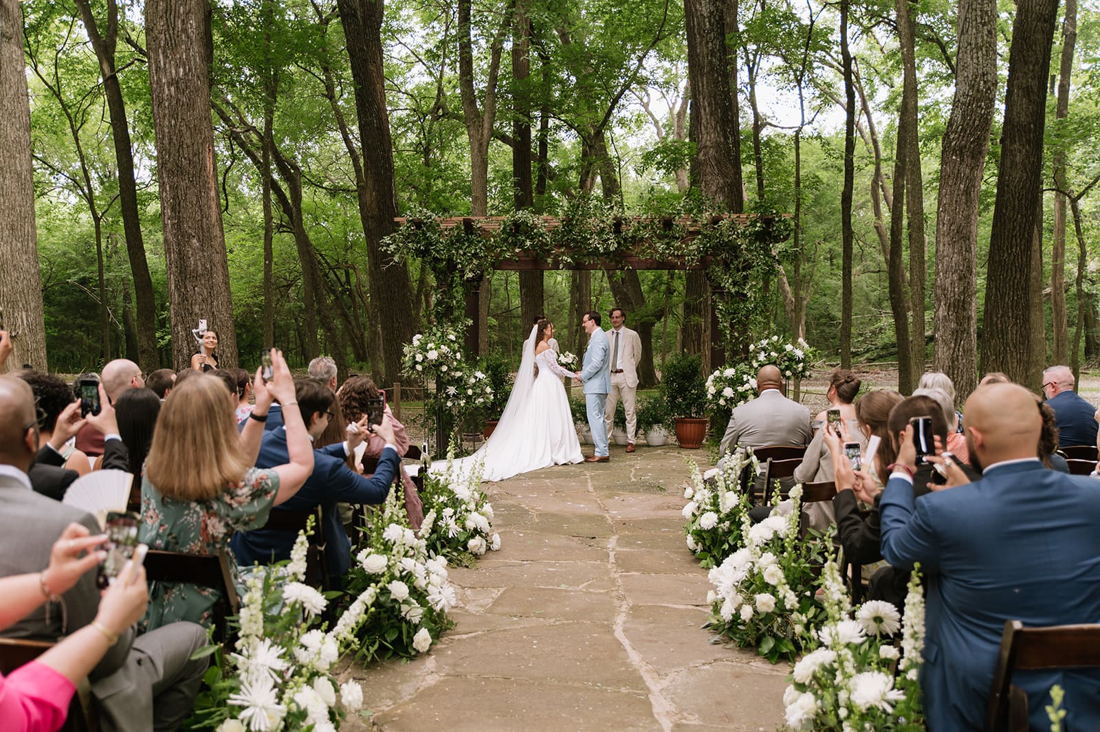 A bride and groom stand together at an outdoor wedding ceremony under a wooden pergola decorated with greenery and flowers, with two men officiating at The Springs Waxahachie