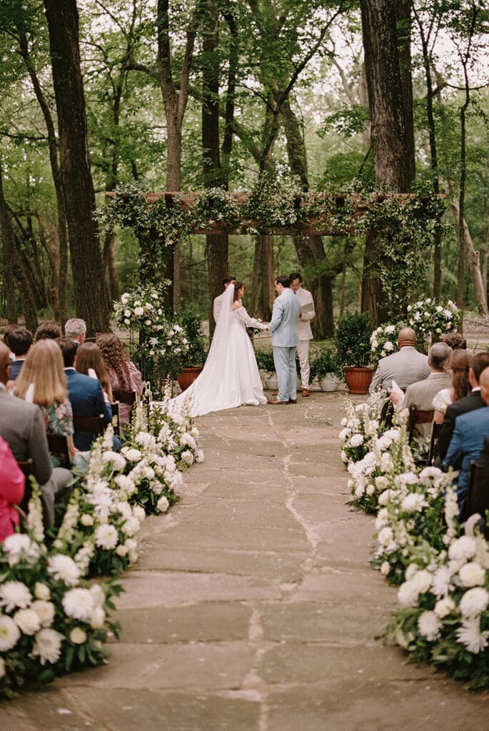 A bride and groom stand together at an outdoor wedding ceremony under a wooden pergola decorated with greenery and flowers, with two men officiating at The Springs Waxahachie
