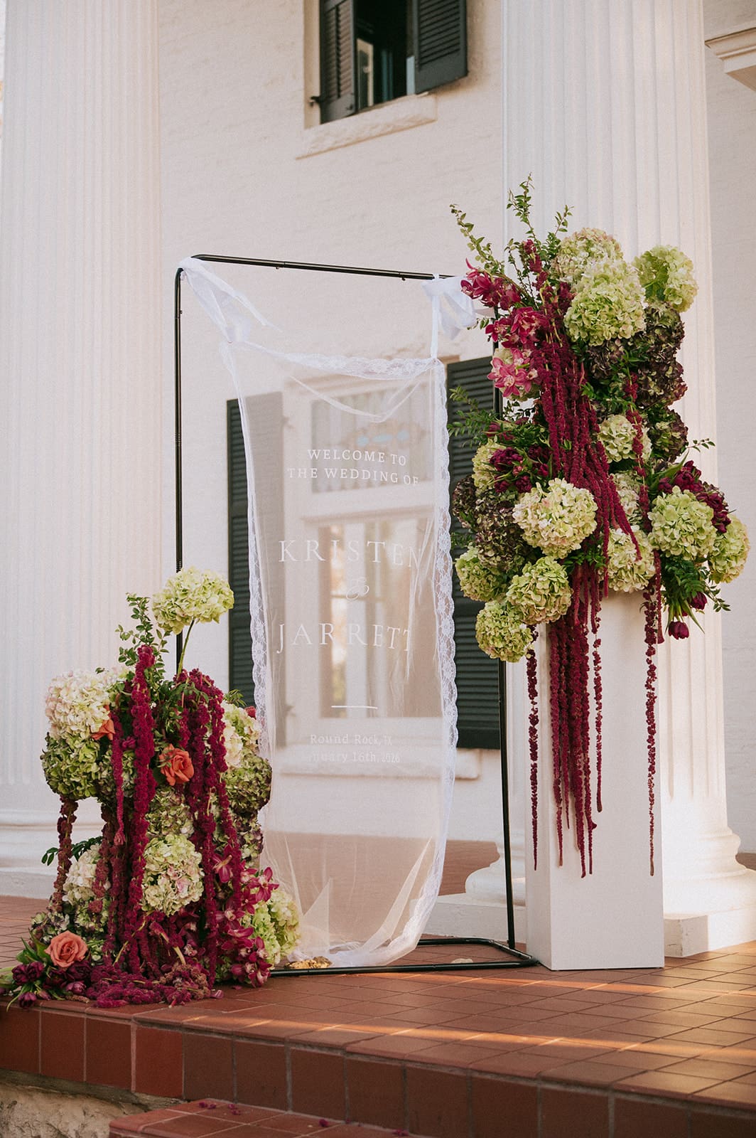 A decorative wedding arch draped with white fabric and adorned with green and burgundy floral arrangements on a brick porch in front of white columns at Woodbine Mansion