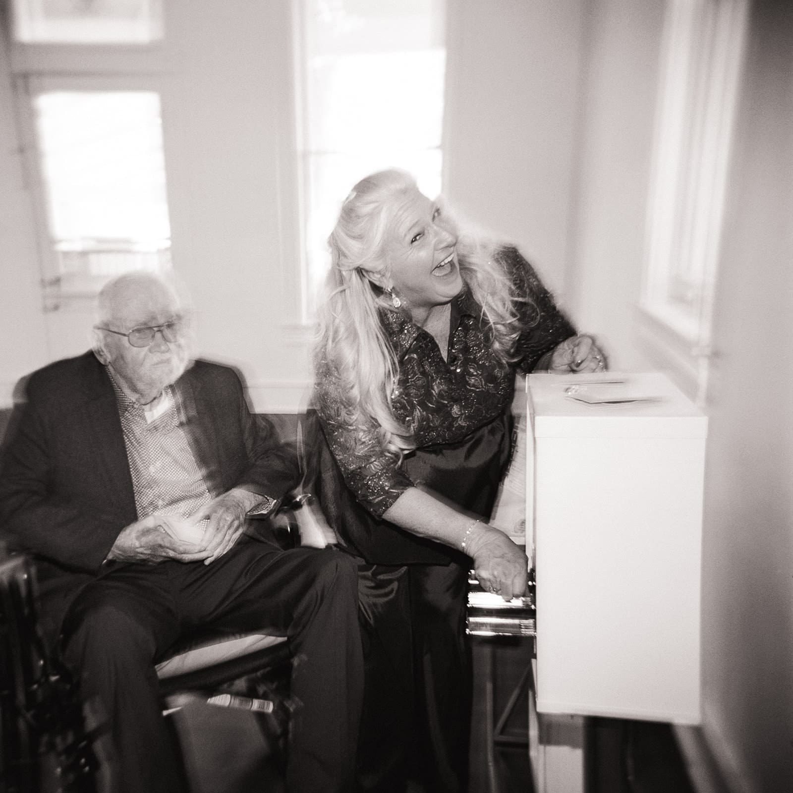 A woman in a dress smiles while turning a handle on a small machine; an older man sits beside her, holding a cup.