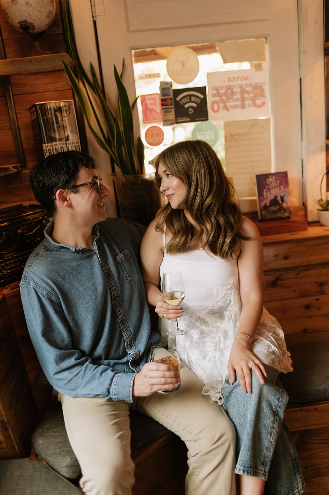 A man and woman sit close together on a bench in a cozy room, holding drinks and smiling at each other for unique engagement photos