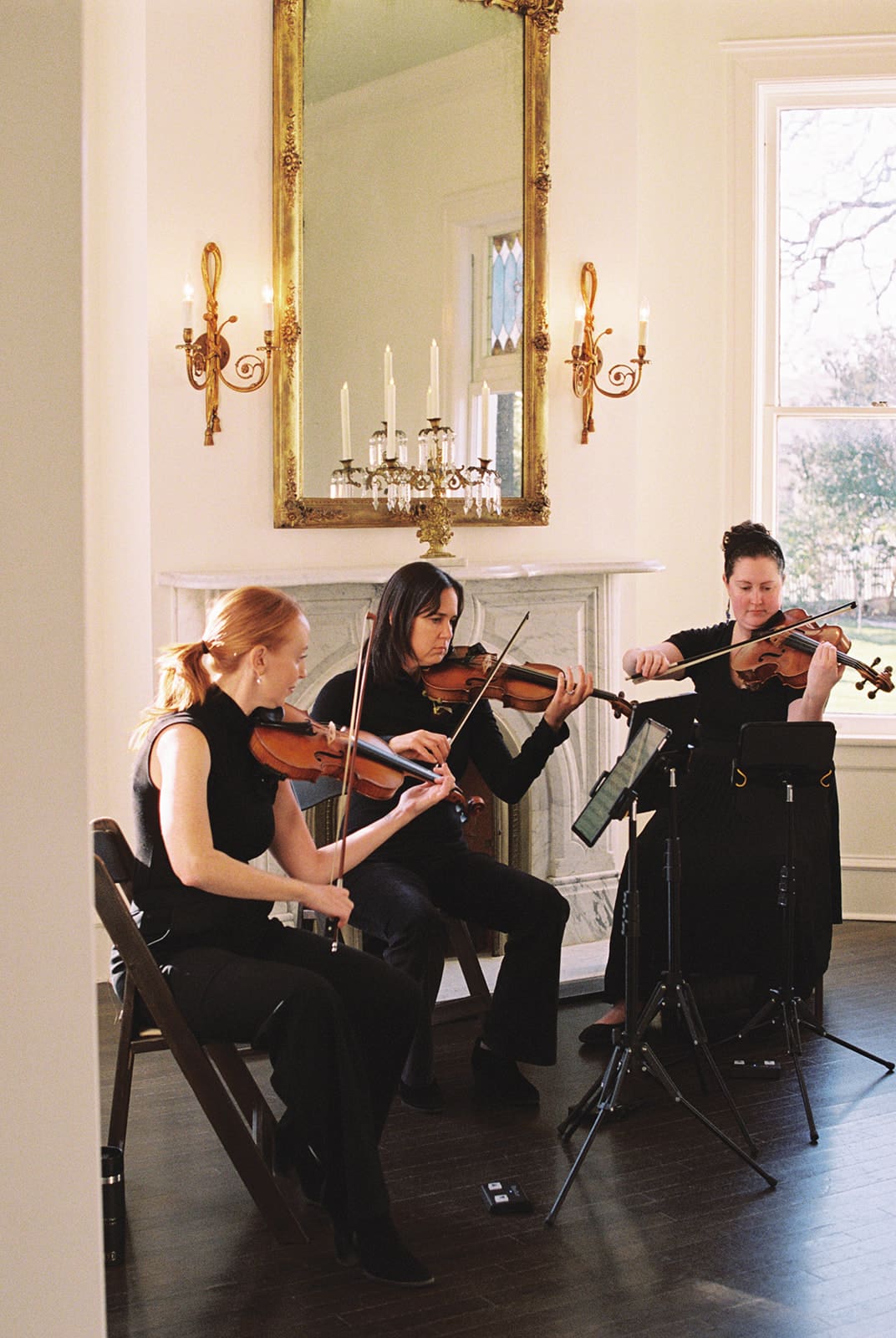 Three women dressed in black perform as a string trio, playing violins and viola in a sunlit room with a fireplace, large mirror, and lit wall sconces.