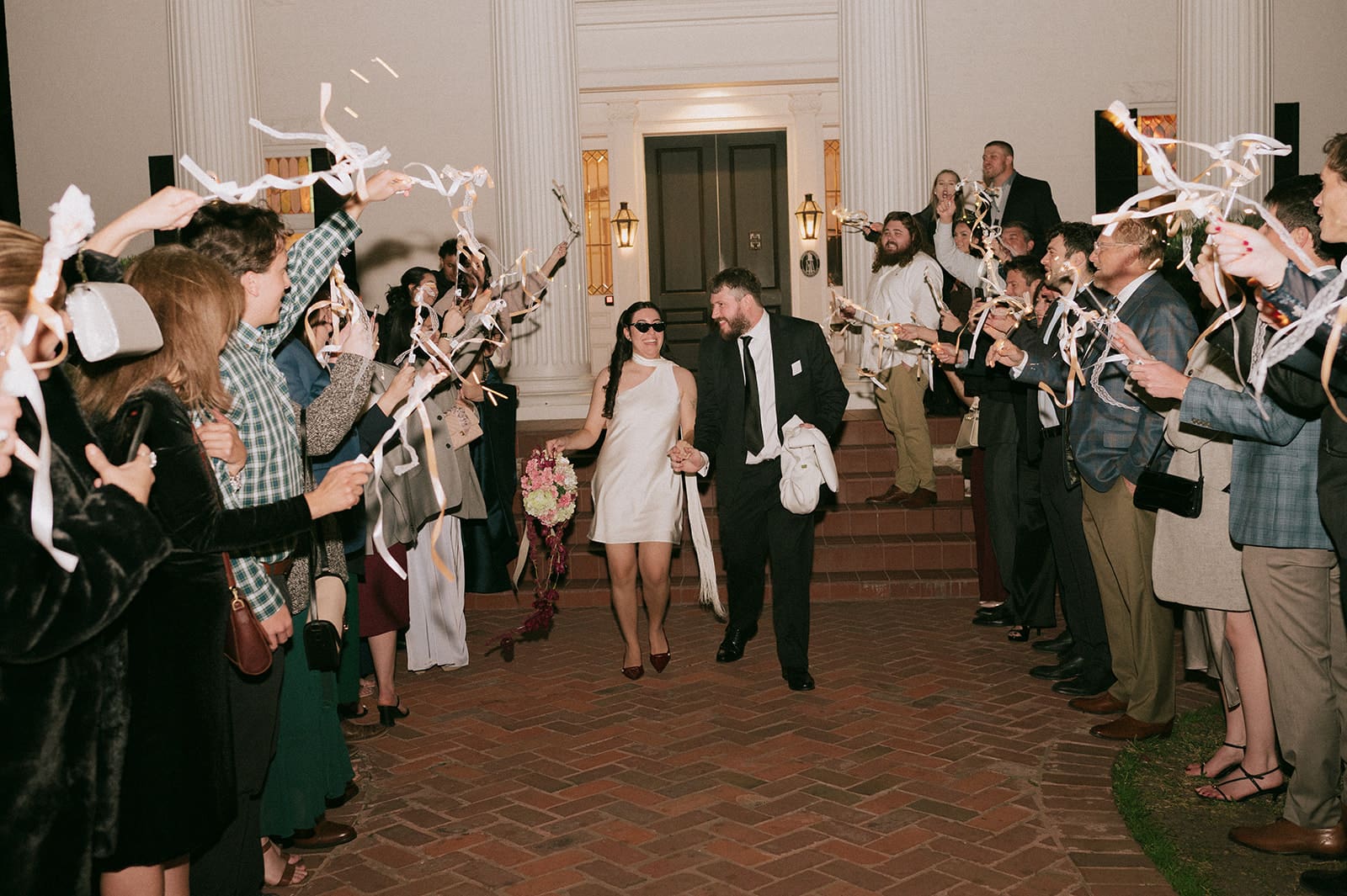 A couple in formal attire walks down steps outside a building while guests on either side cheer and wave streamers.