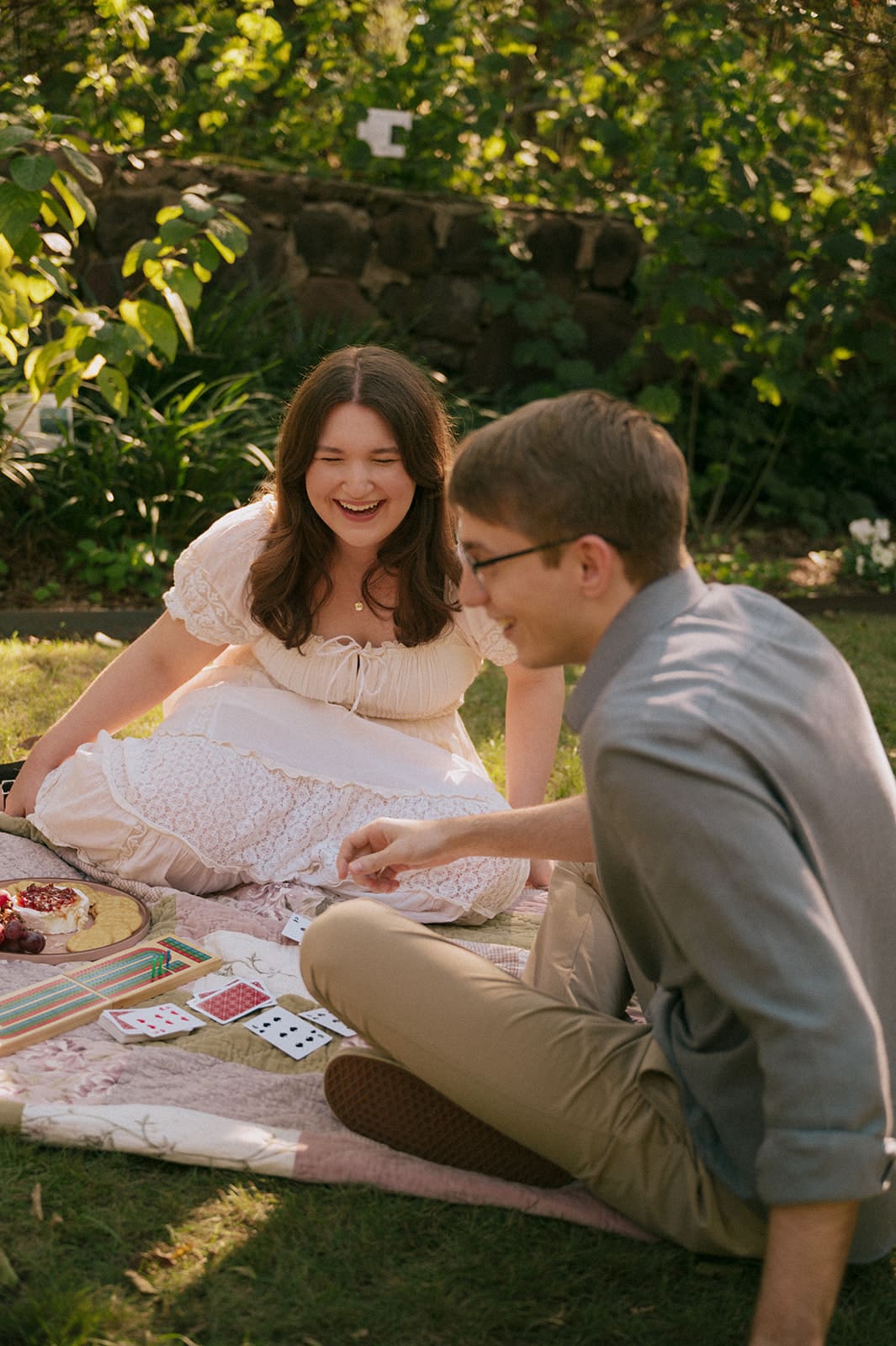 Two people sit on a blanket outdoors, playing cards and smiling, surrounded by greenery and snacks on a sunny day for unique engagement photos
