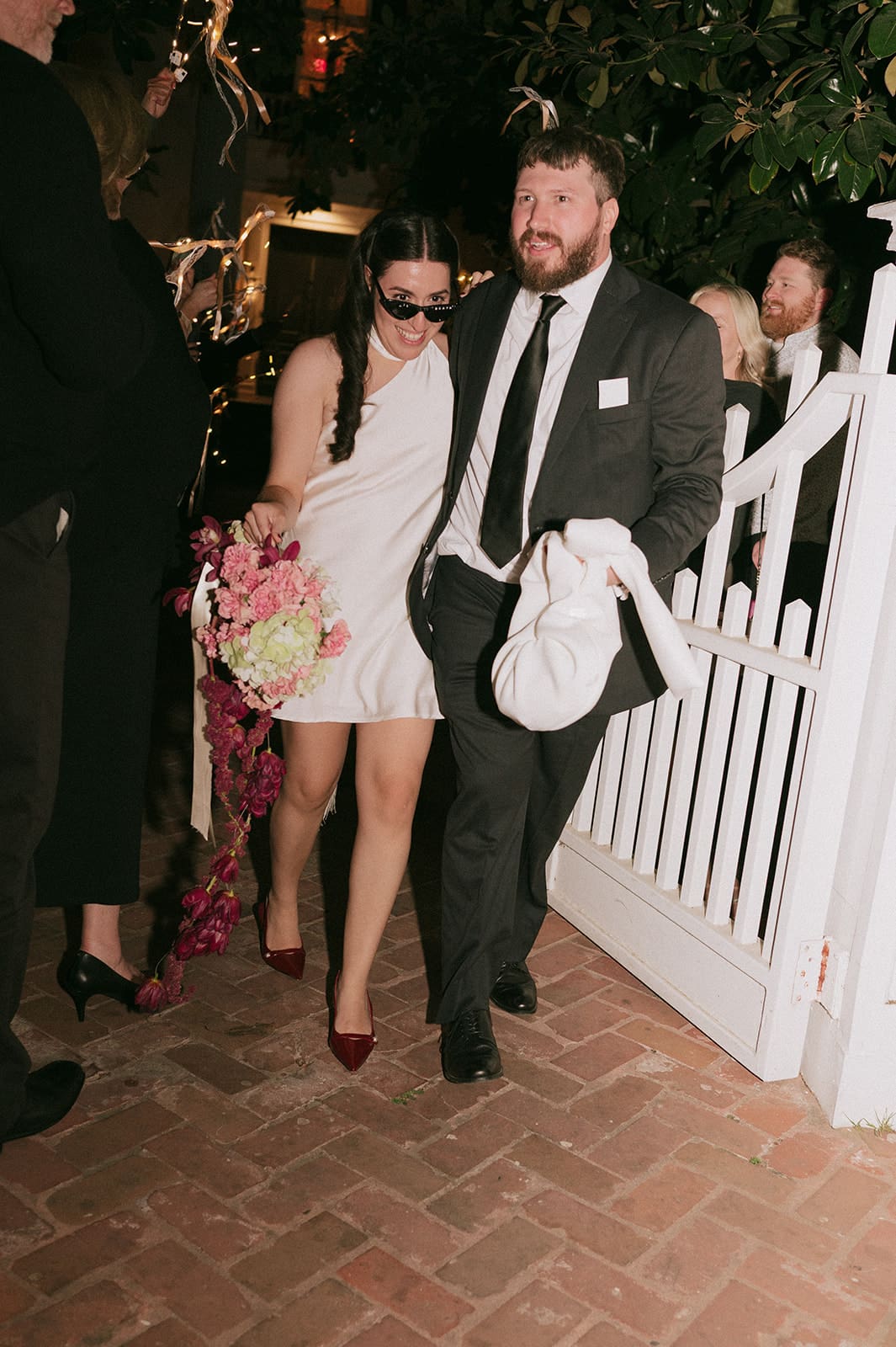 A woman in a white dress holding a bouquet walks beside a man in a suit through a white gate at night.