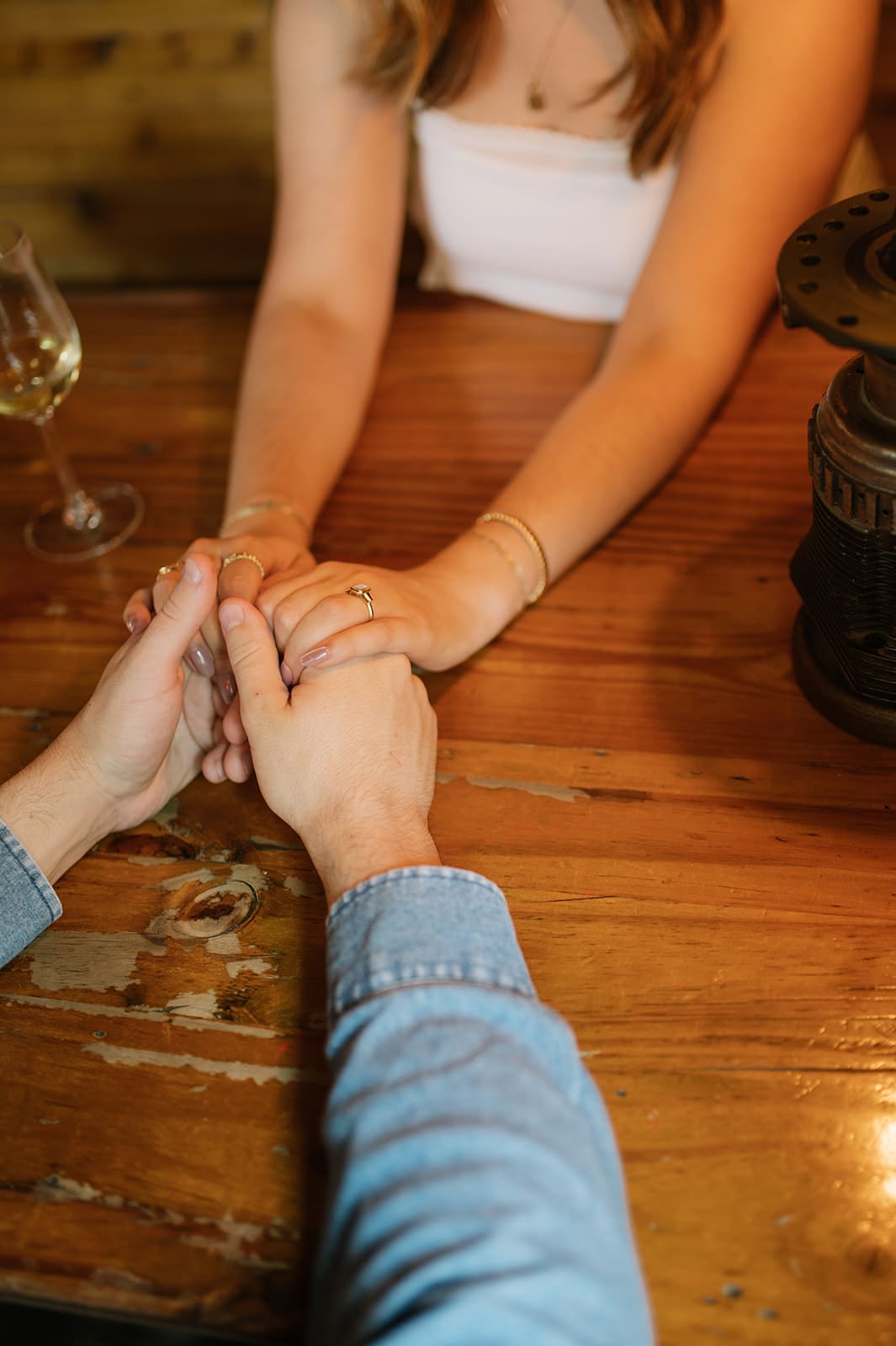 Two people sit across from each other at a wooden table, holding hands. One person wears a white top, the other has a blue long-sleeve shirt. A glass of wine is on the table.