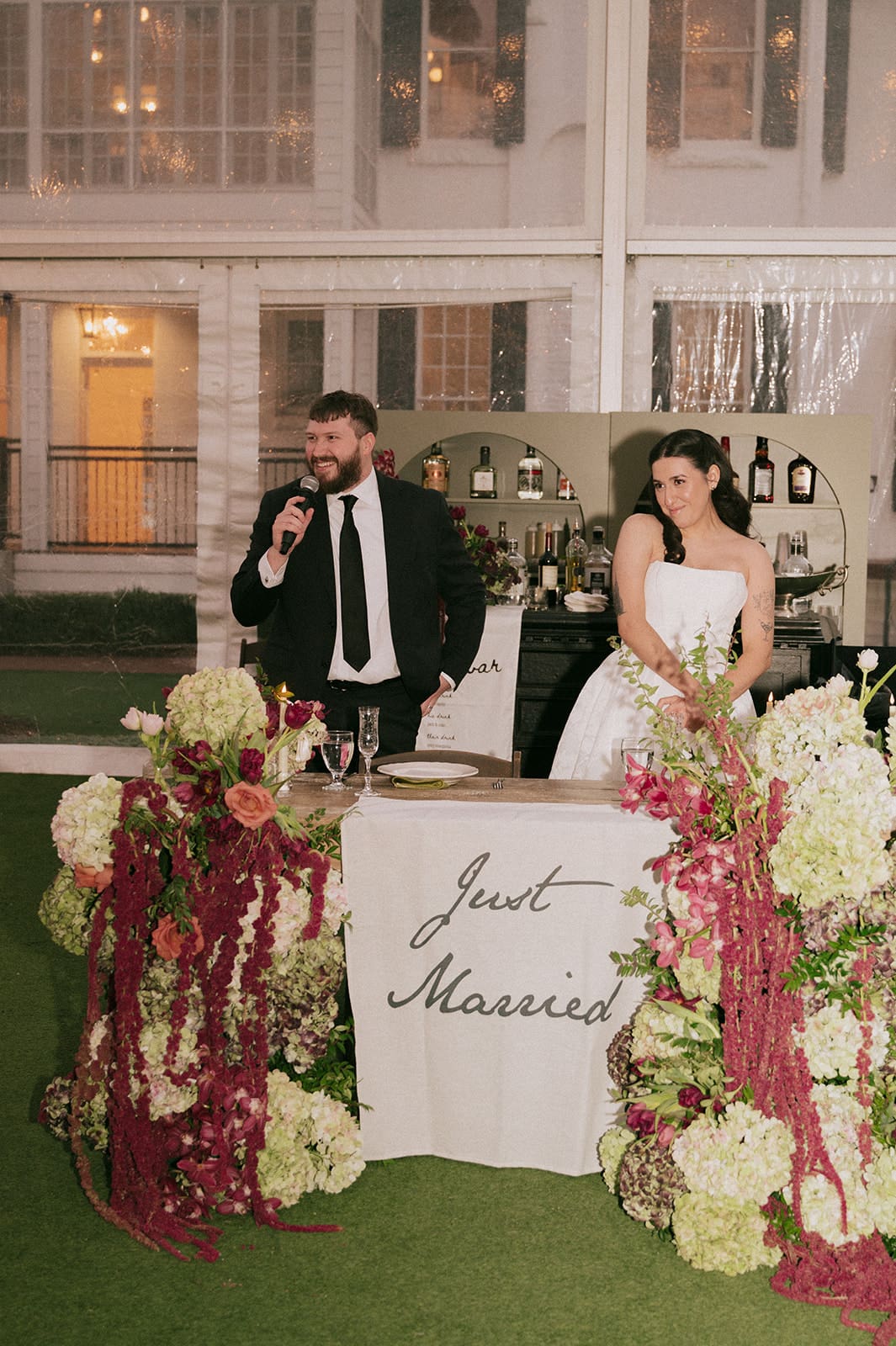 A man in a suit speaks into a microphone next to a woman in a white wedding dress standing behind a table with a "Just Married" sign and floral arrangements.
