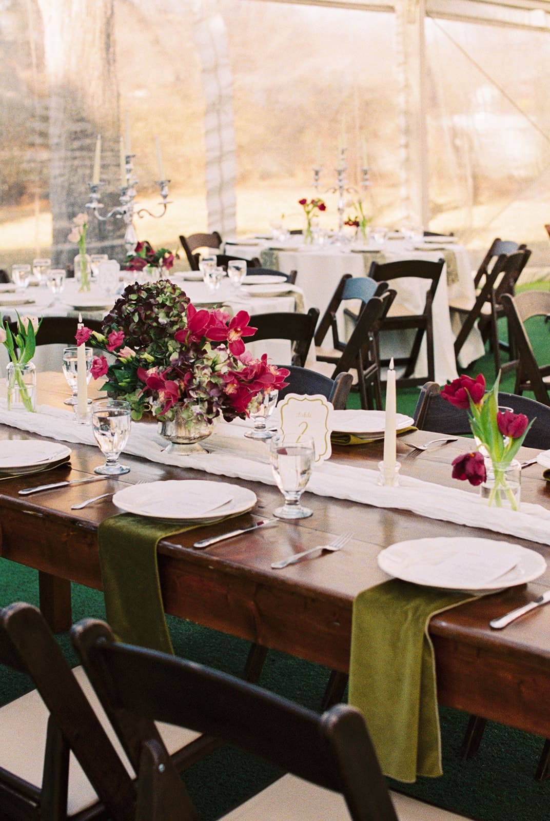 A wooden table set with white plates, glassware, green napkins, and floral centerpieces, surrounded by black chairs in an outdoor, tented setting at Woodbine Mansion