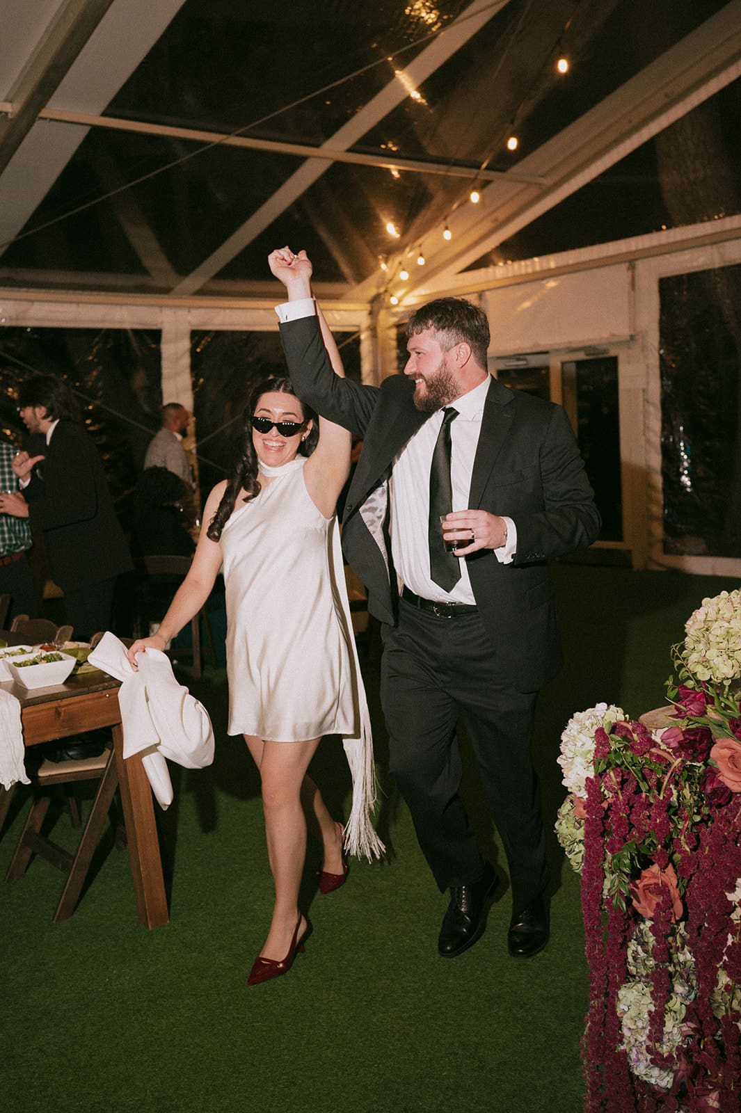 A woman in a white dress and sunglasses walks hand-in-hand with a man in a suit, both smiling, at an indoor event with string lights overhead at Woodbine Mansion