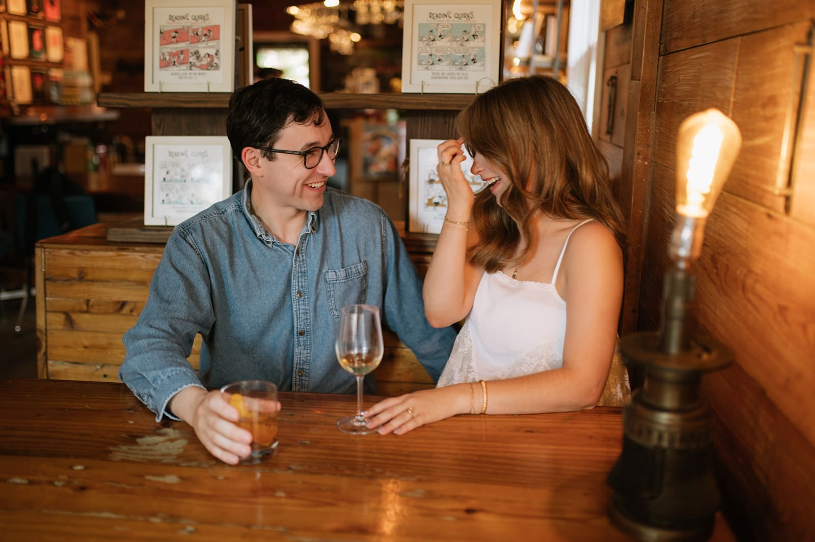 A man and a woman sit at a wooden table with drinks, smiling and talking in a cozy, warmly lit setting.