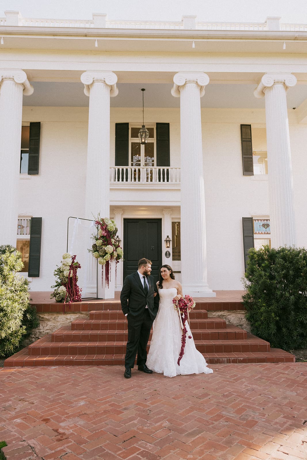 A man in a black suit and a woman in a white wedding dress stand side by side outdoors in front of the woodbine mansion