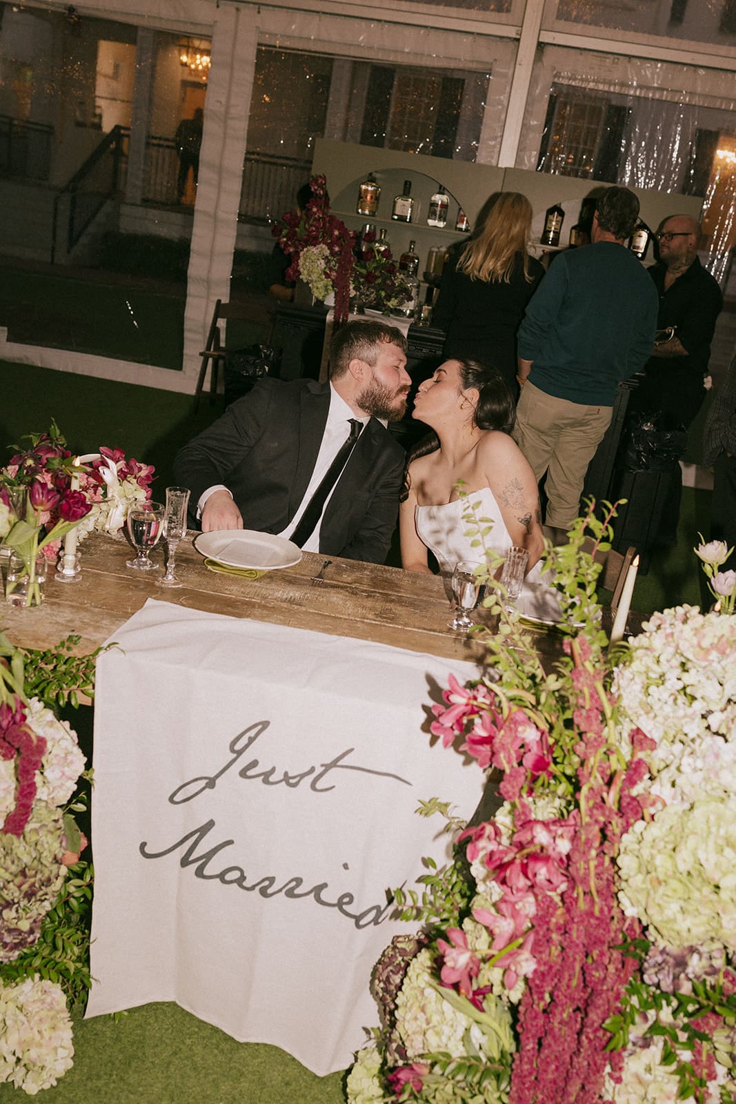 A man in a suit speaks into a microphone next to a woman in a white wedding dress standing behind a table with a "Just Married" sign and floral arrangements.