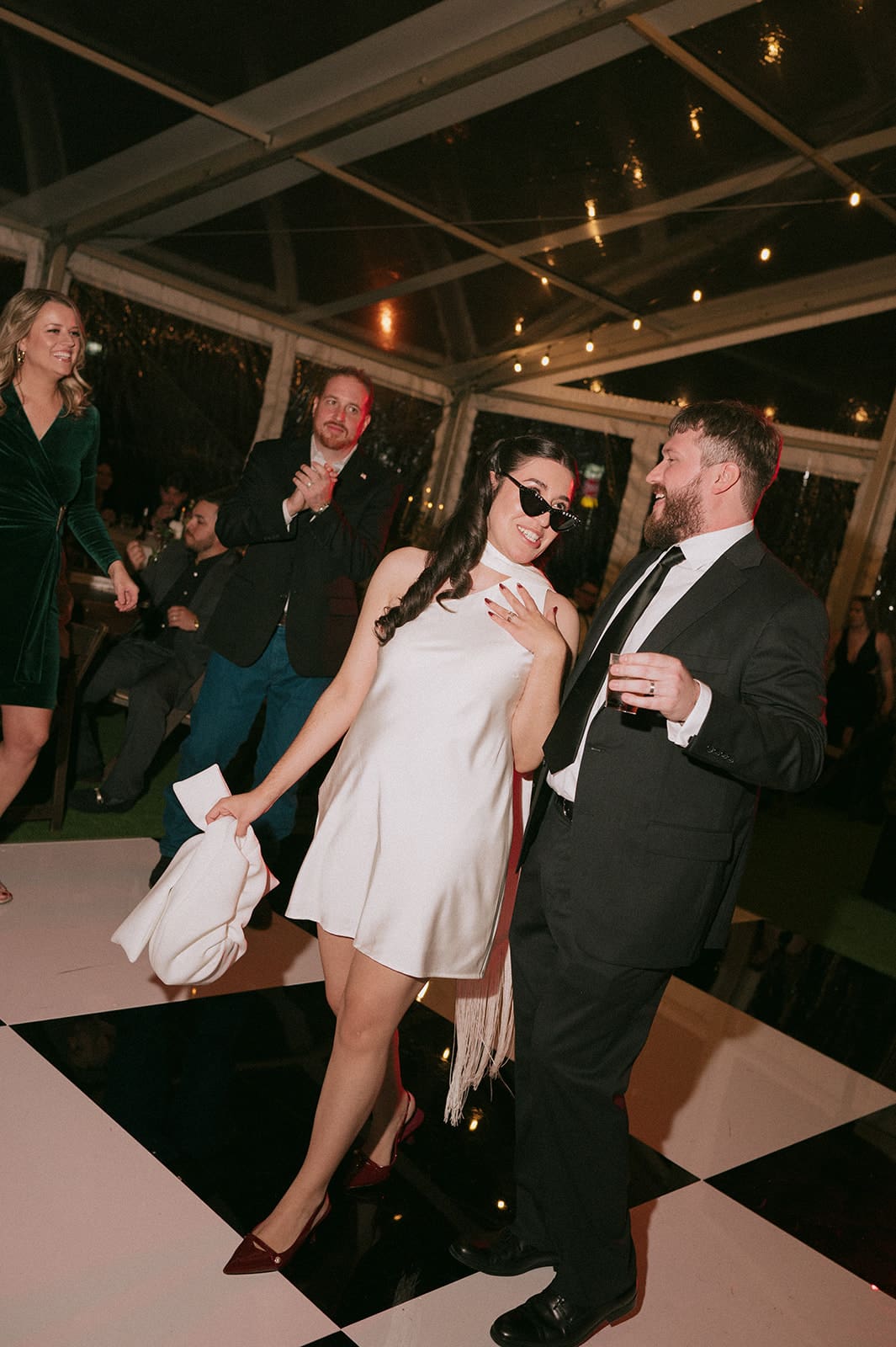A woman in a white dress and sunglasses walks hand-in-hand with a man in a suit, both smiling, at an indoor event with string lights overhead at Woodbine Mansion