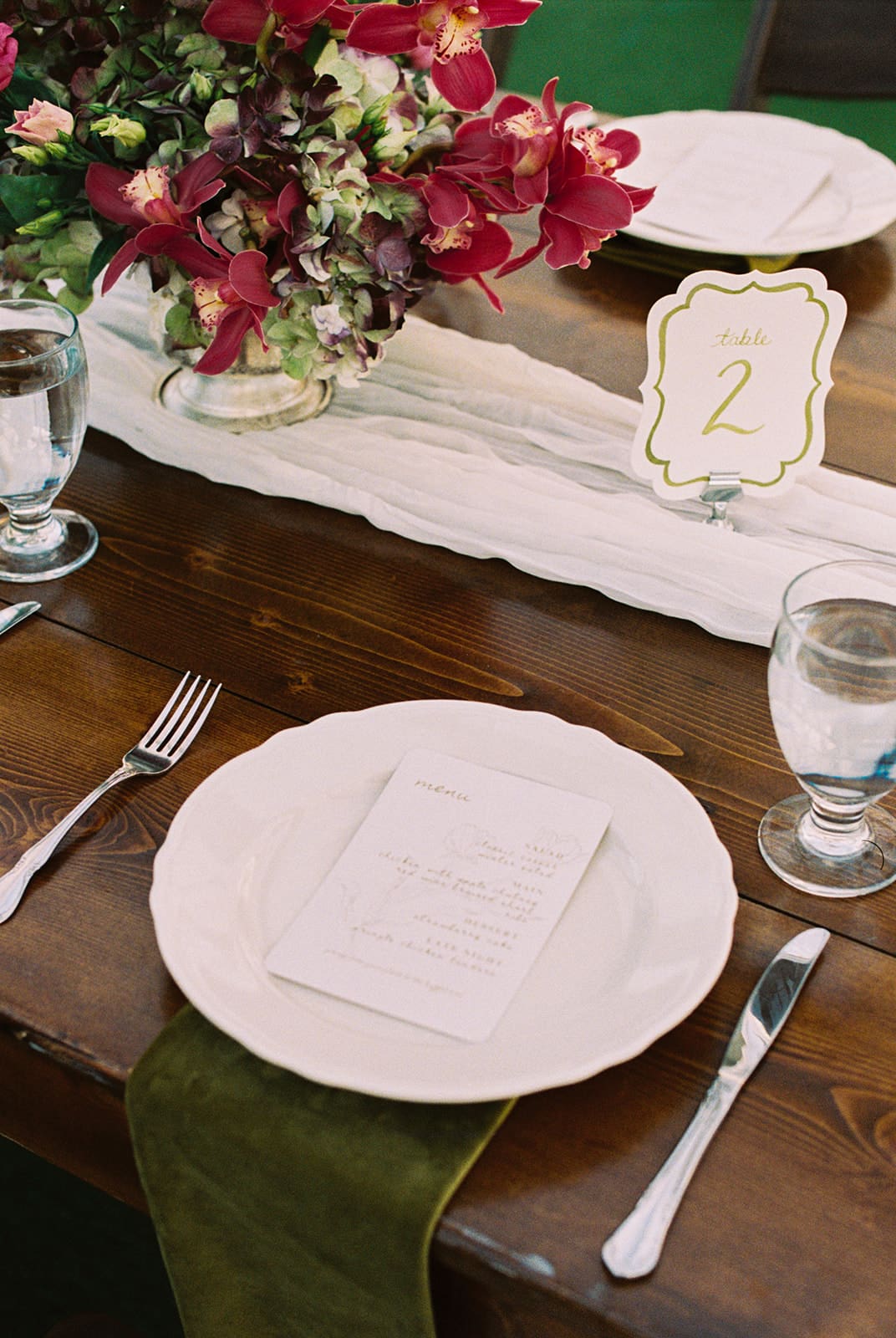 A place setting with a white plate, menu, cutlery, two glasses of water, a green napkin, floral centerpiece, and a table number card reading “2” on a wooden table at Woodbine Mansion