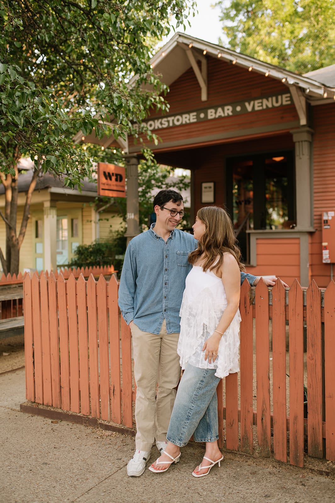 A couple stands close together, smiling at each other in front of a small building with a sign that reads “BOOKSTORE • BAR • VENUE,” surrounded by a red fence and trees.