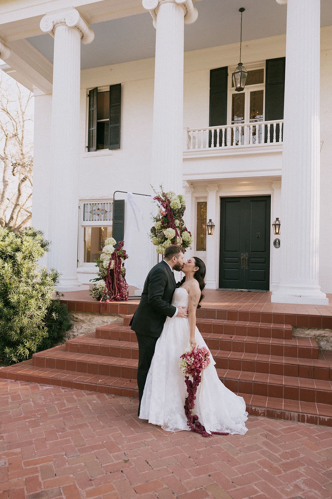 A man in a black suit and a woman in a white wedding dress stand side by side outdoors in front of the woodbine mansion