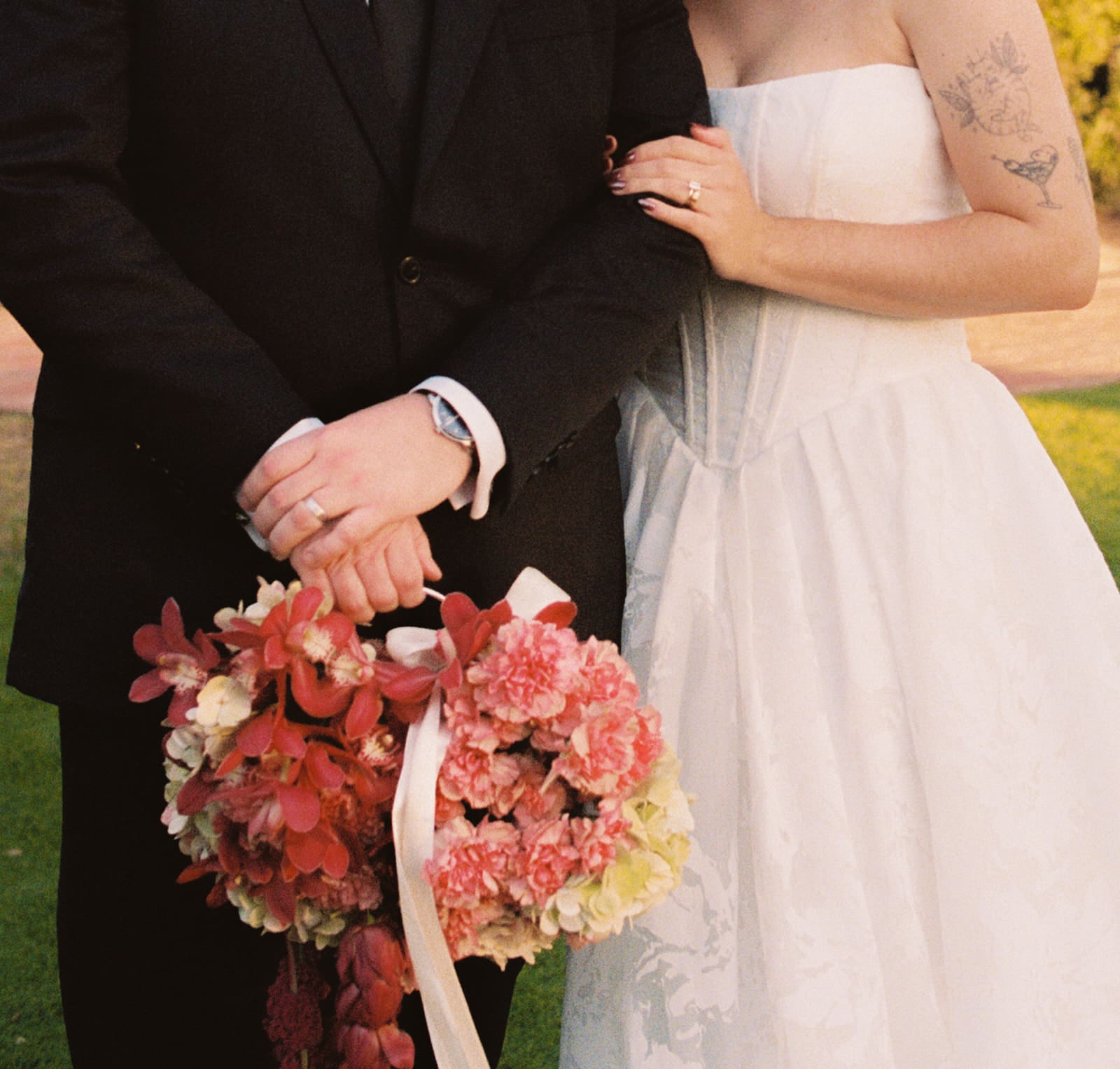 A bride in a white dress and a groom in a black suit stand together outside, holding a bouquet of pink and white flowers.