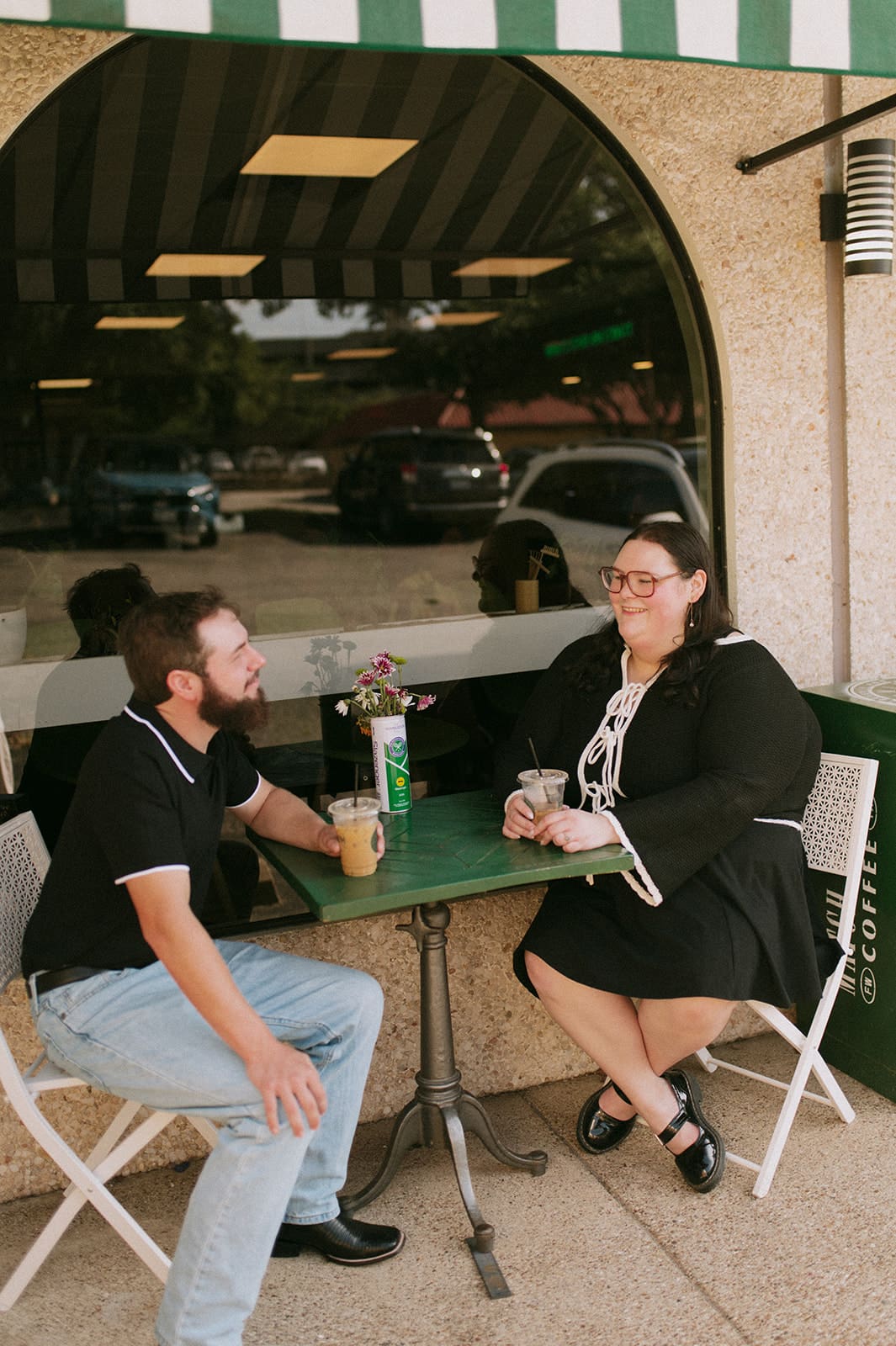 Two people sit at a small table outside Match Point Coffee, a café with striped green and white awnings and a large sign above the entrance.