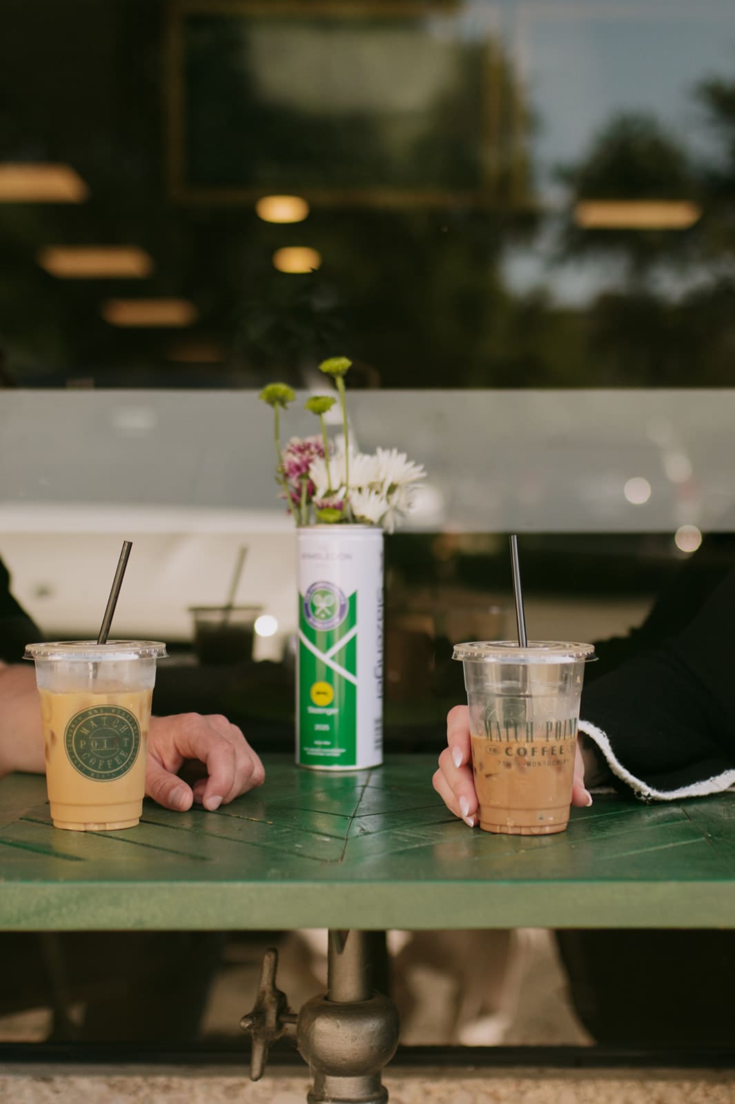 Two people sit across from each other at a green table with iced coffee drinks; a small vase with flowers is placed between them.