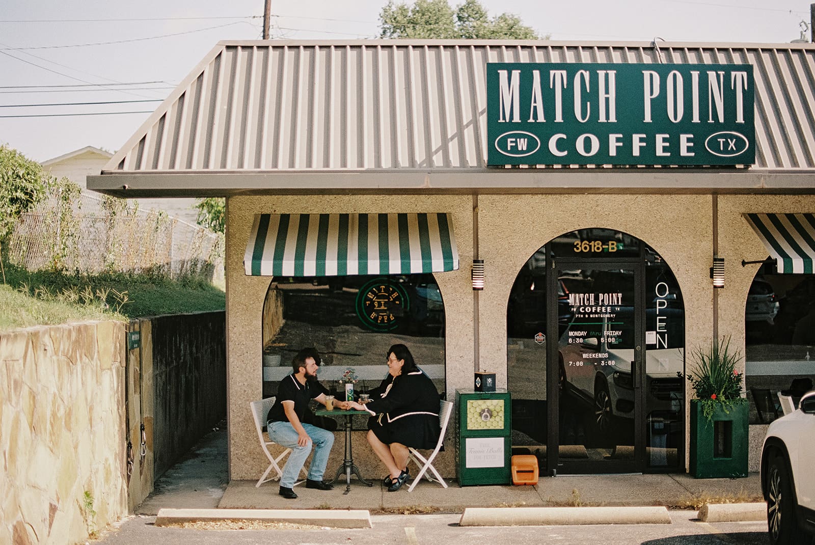 Two people sit at a small table outside Match Point Coffee, a café with striped green and white awnings and a large sign above the entrance.