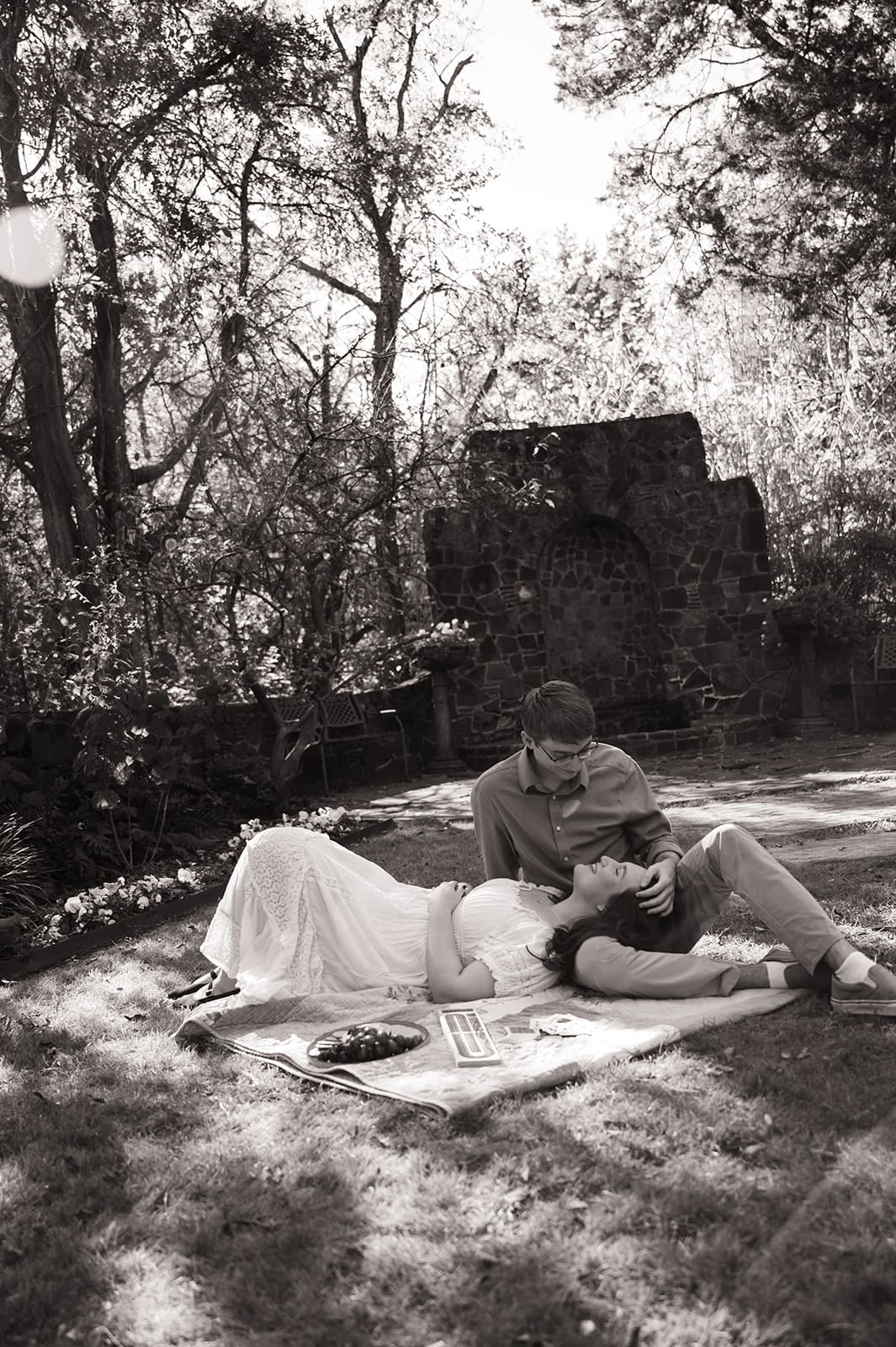 Two people sit on a blanket outdoors, playing cards and smiling, surrounded by greenery and snacks on a sunny day for unique engagement photos