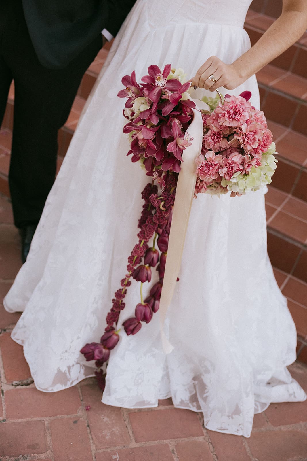 A person in a white wedding dress holding a bouquet of pink and purple flowers stands next to another person in dark formal attire on brick steps.