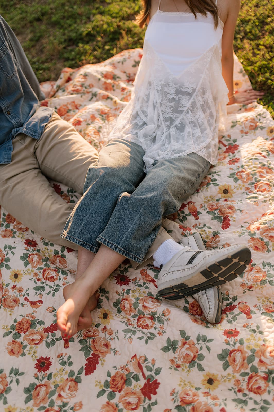 Two people sit on a floral-patterned blanket on grass, with their legs extended and partially overlapping. One person is barefoot, the other wears sneakers for unique engagement pictures