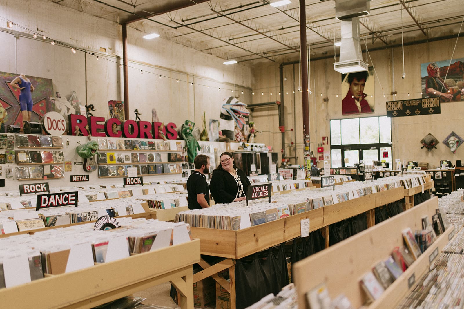 Interior of a record store with rows of vinyl records, labeled sections, and two people standing near the center. Decor and posters are visible on the walls for unique engagement pictures