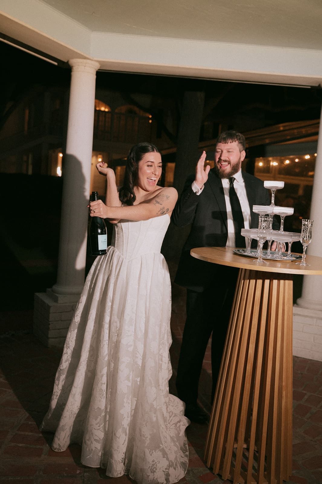 A bride in a white gown holds a bottle while standing next to a groom in a suit near a champagne tower, both smiling and gesturing joyfully.