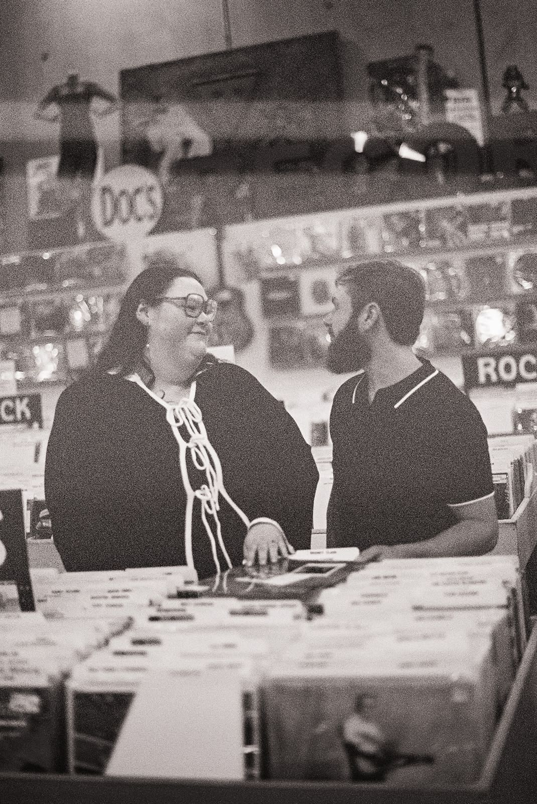 Interior of a record store with rows of vinyl records, labeled sections, and two people standing near the center. Decor and posters are visible on the walls for unique engagement pictures