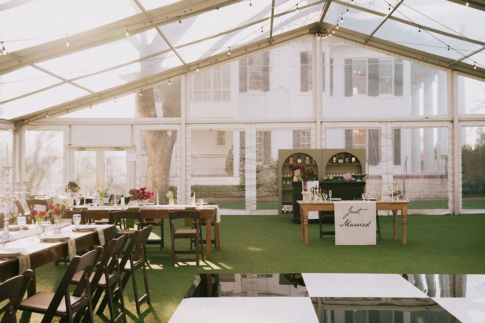 A clear tent set up for an event with tables, chairs, and a bar inside, in front of a large white house with visible greenery outside.