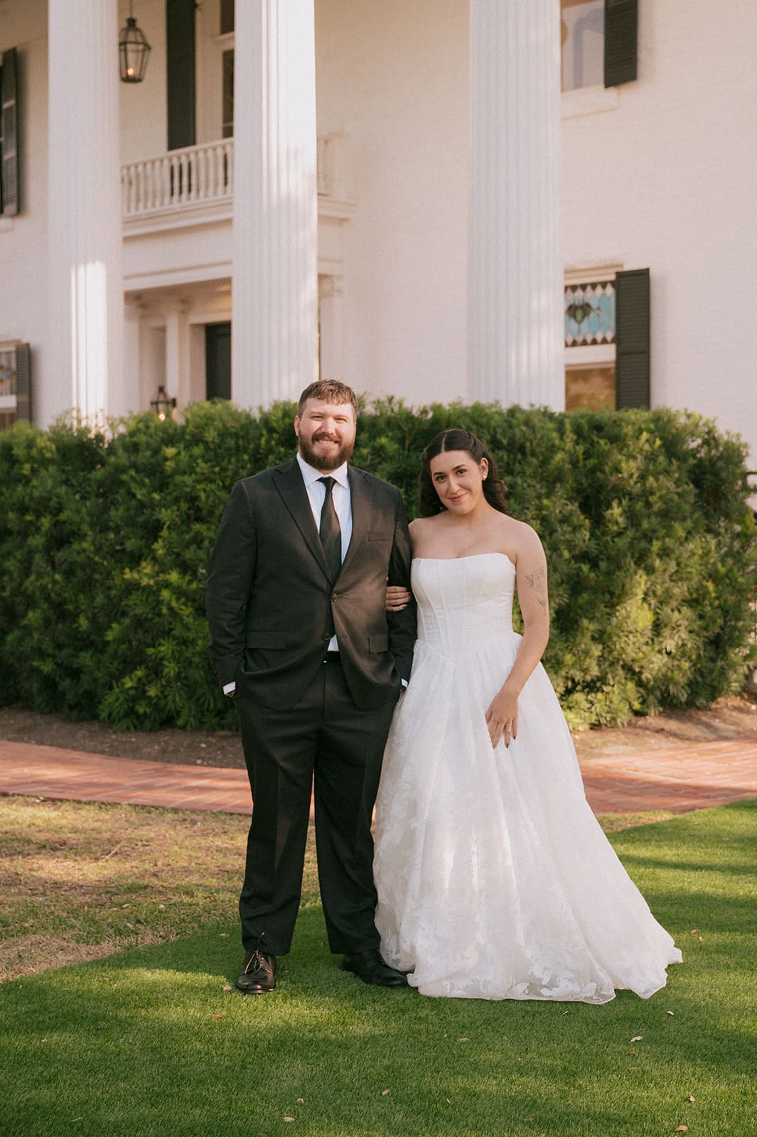 A man in a black suit and a woman in a white wedding dress stand side by side outdoors in front of the woodbine mansion