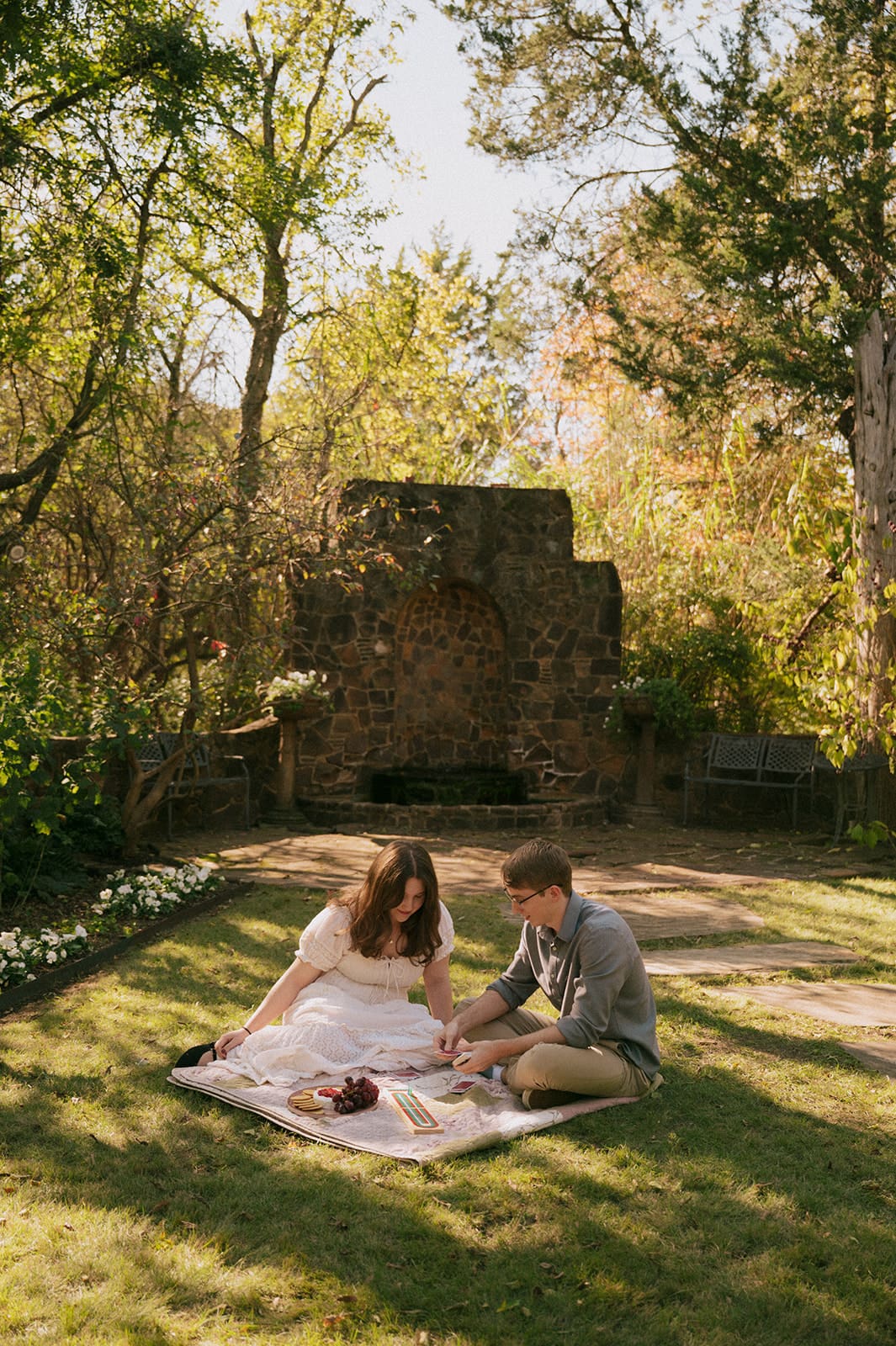 Two people sit on a blanket outdoors, playing cards and smiling, surrounded by greenery and snacks on a sunny day for unique engagement photos