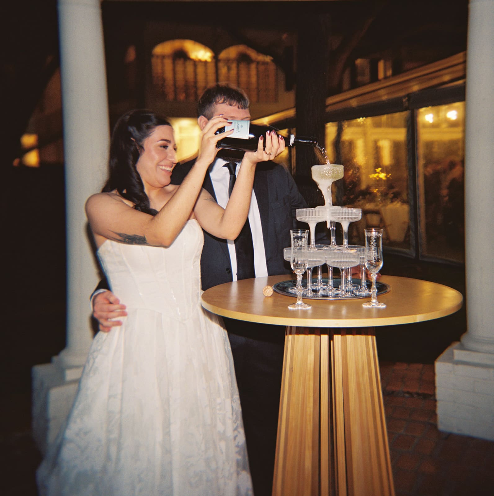 A bride in a white gown holds a bottle while standing next to a groom in a suit near a champagne tower, both smiling and gesturing joyfully.