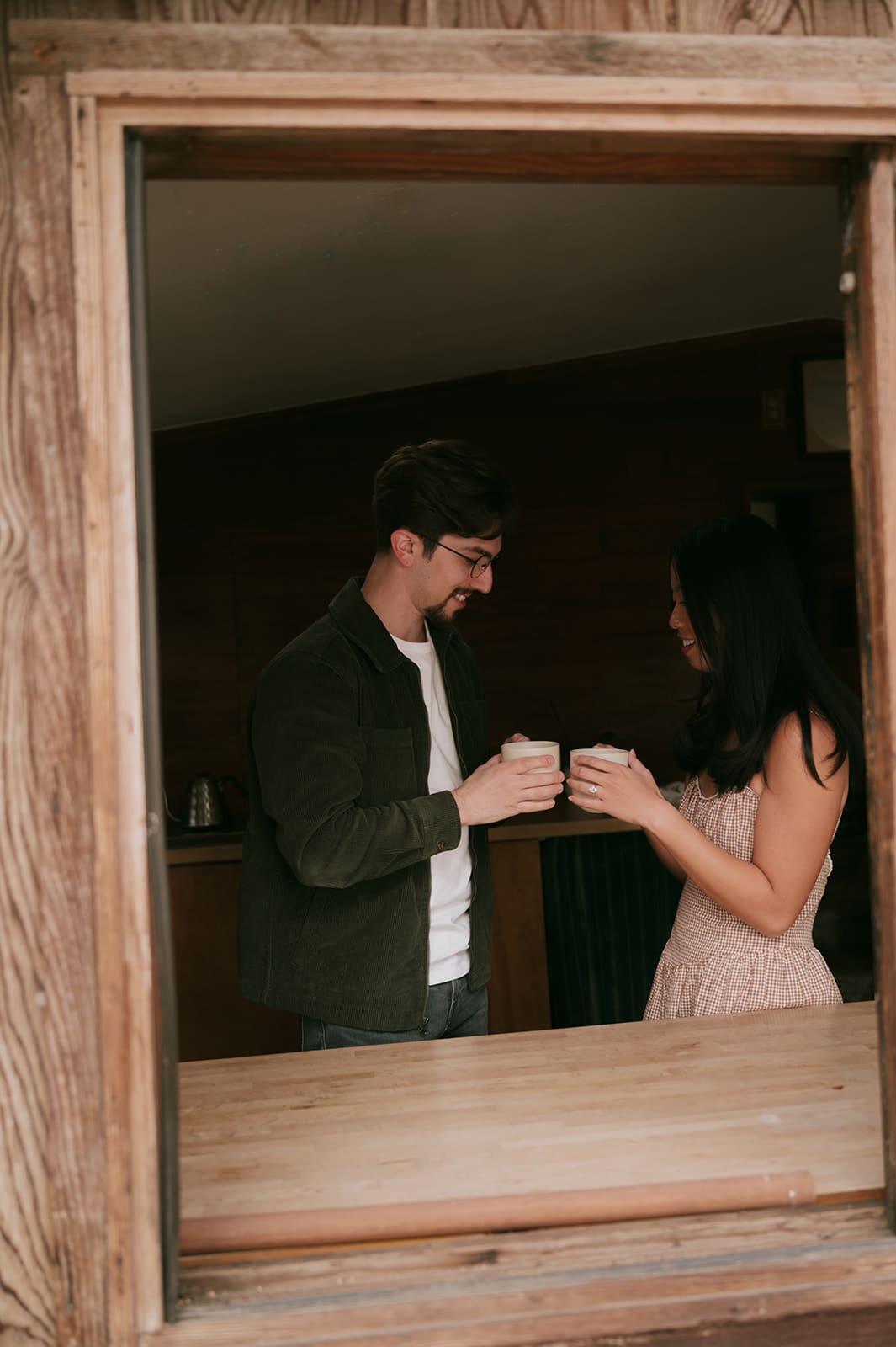 A man and a woman stand together by an open window, facing each other and holding mugs inside a wooden cabin.