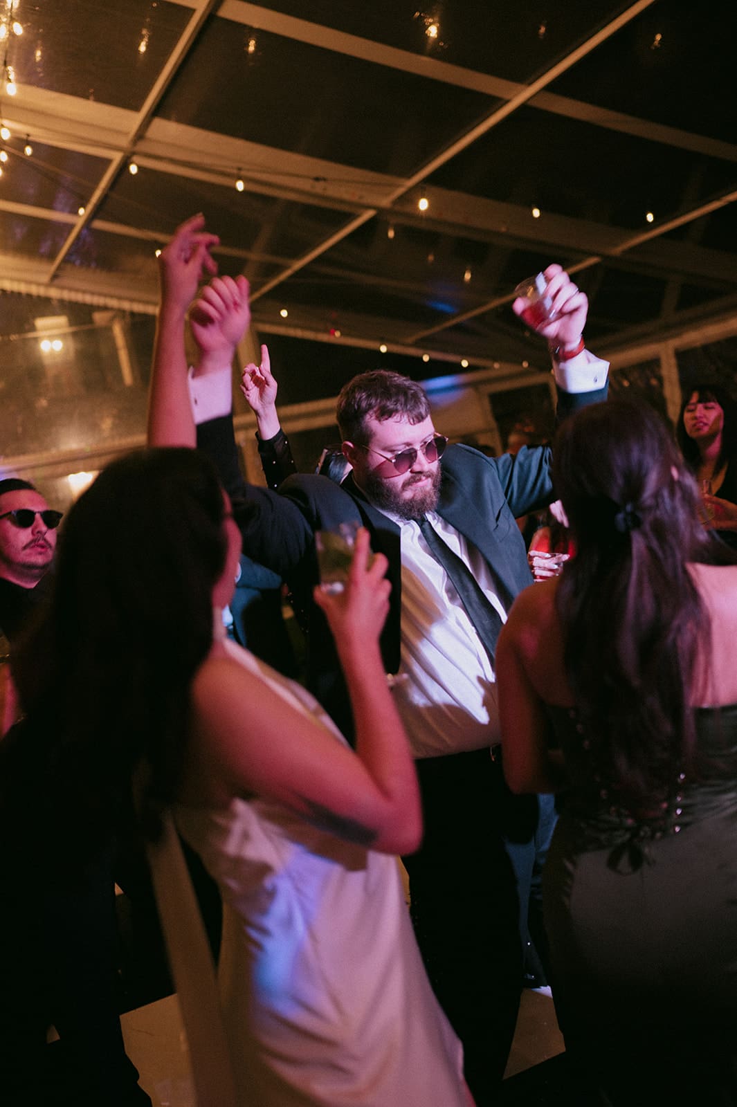 A group of people dressed in formal attire dance and raise their hands at a nighttime indoor event with string lights overhead.