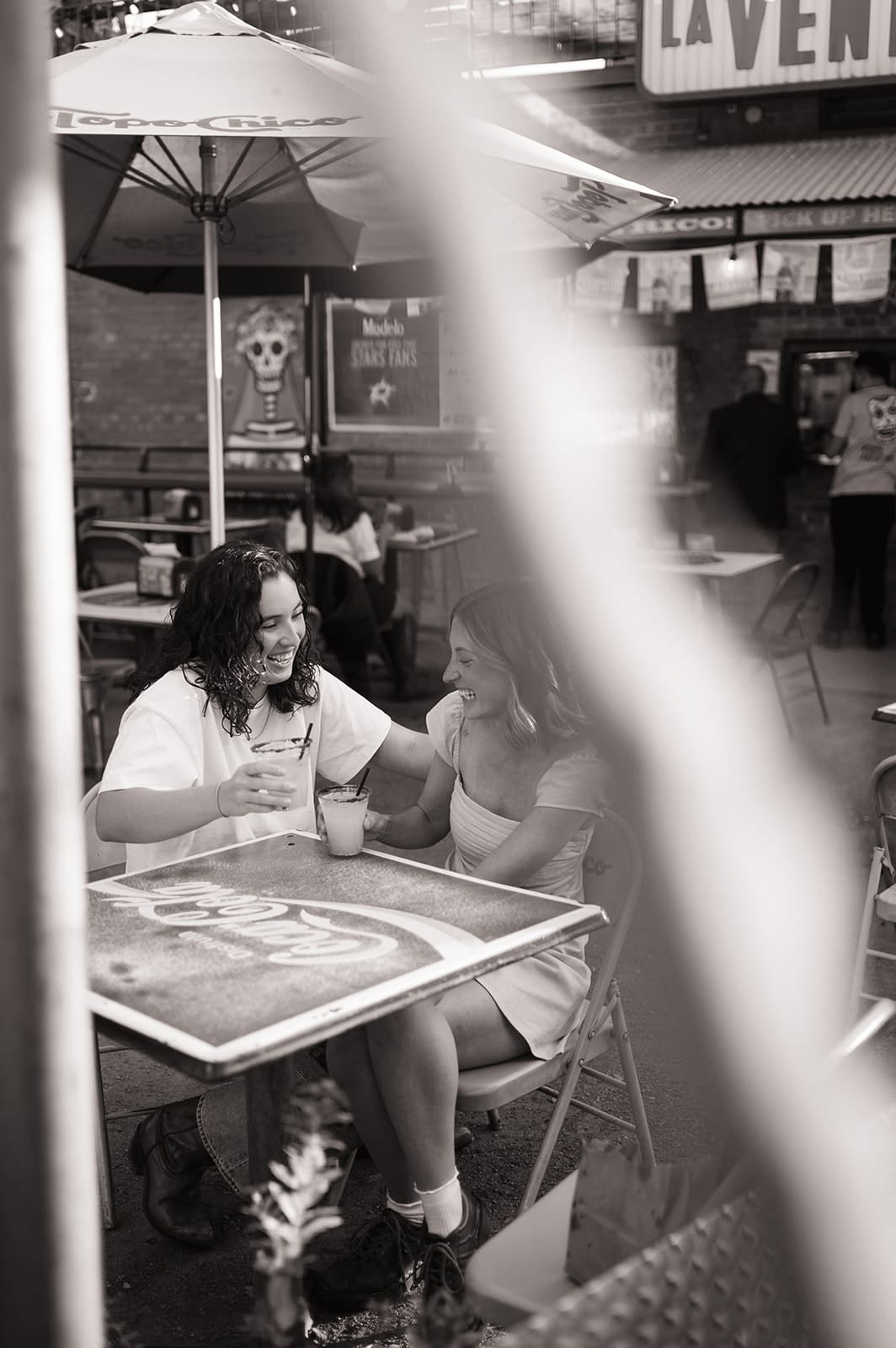 Two women sit at a wooden table under a yellow umbrella, sharing a drink with straws and smiling at each other in an outdoor setting with colorful murals.