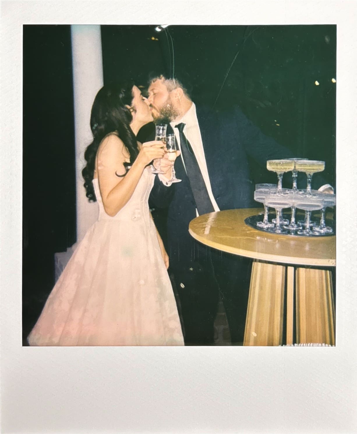 A bride in a white gown holds a bottle while standing next to a groom in a suit near a champagne tower, both smiling and gesturing joyfully.