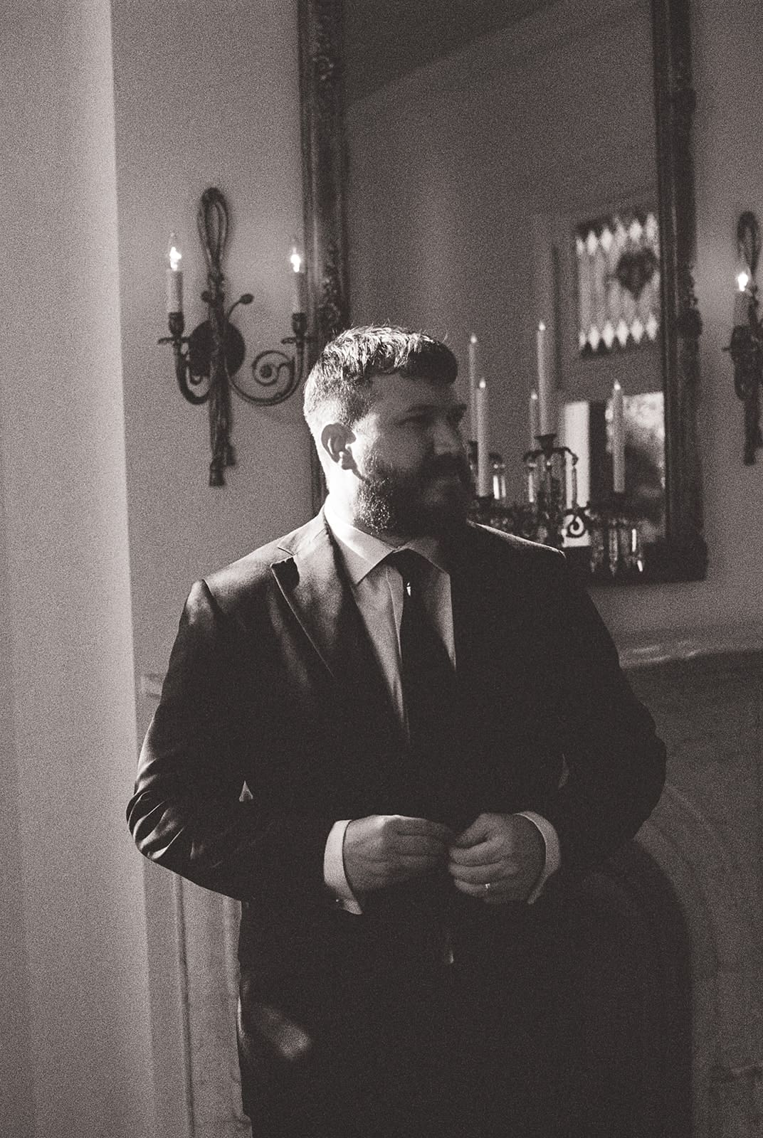 A man in a suit stands indoors near a fireplace and a large mirror, adjusting his jacket. The image is in black and white.
