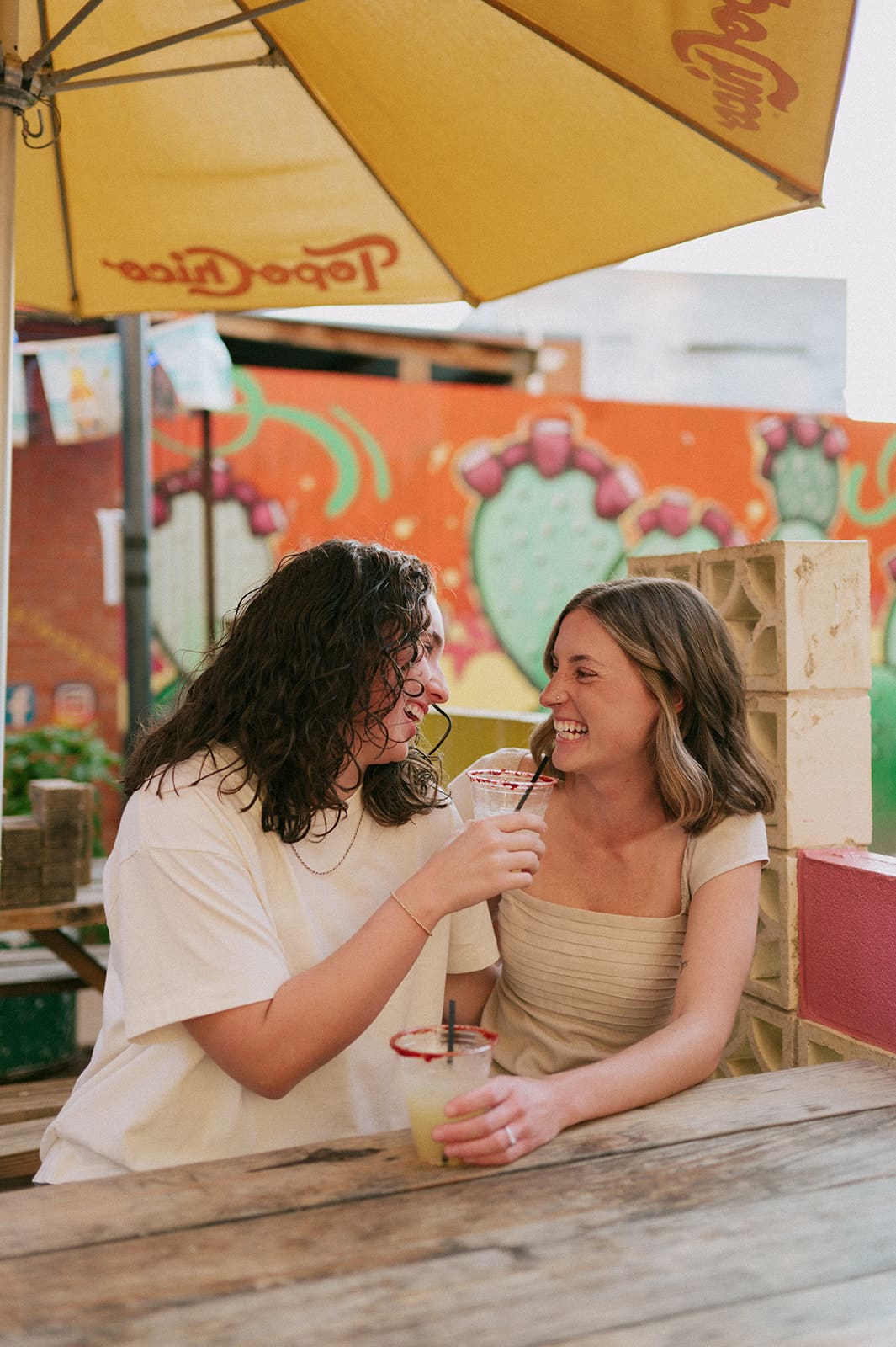Two women sit at a wooden table under a yellow umbrella, sharing a drink with straws and smiling at each other in an outdoor setting with colorful murals.