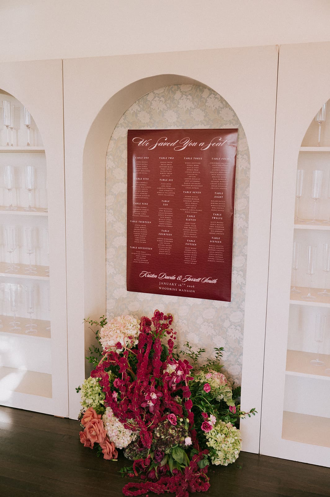 A maroon seating chart with white text is displayed on a white arched wall, with an arrangement of pink and red flowers at the base at Woodbine Mansion